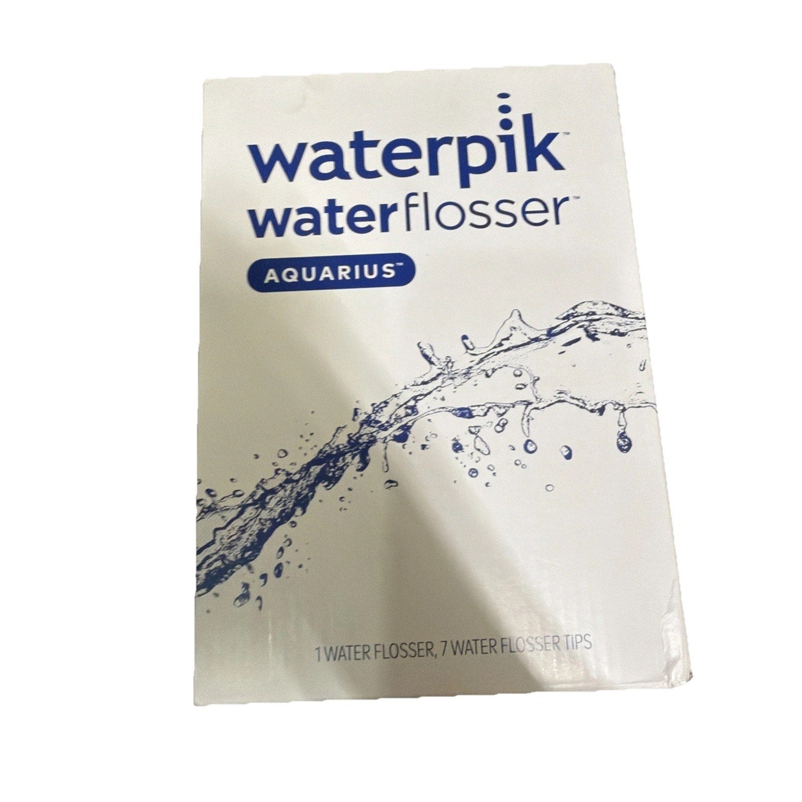 Front view of Waterpik Water Flosser Aquarius packaging featuring the brand name, a blue water splash graphic, and text indicating the inclusion of one flosser and seven tips on a white background.