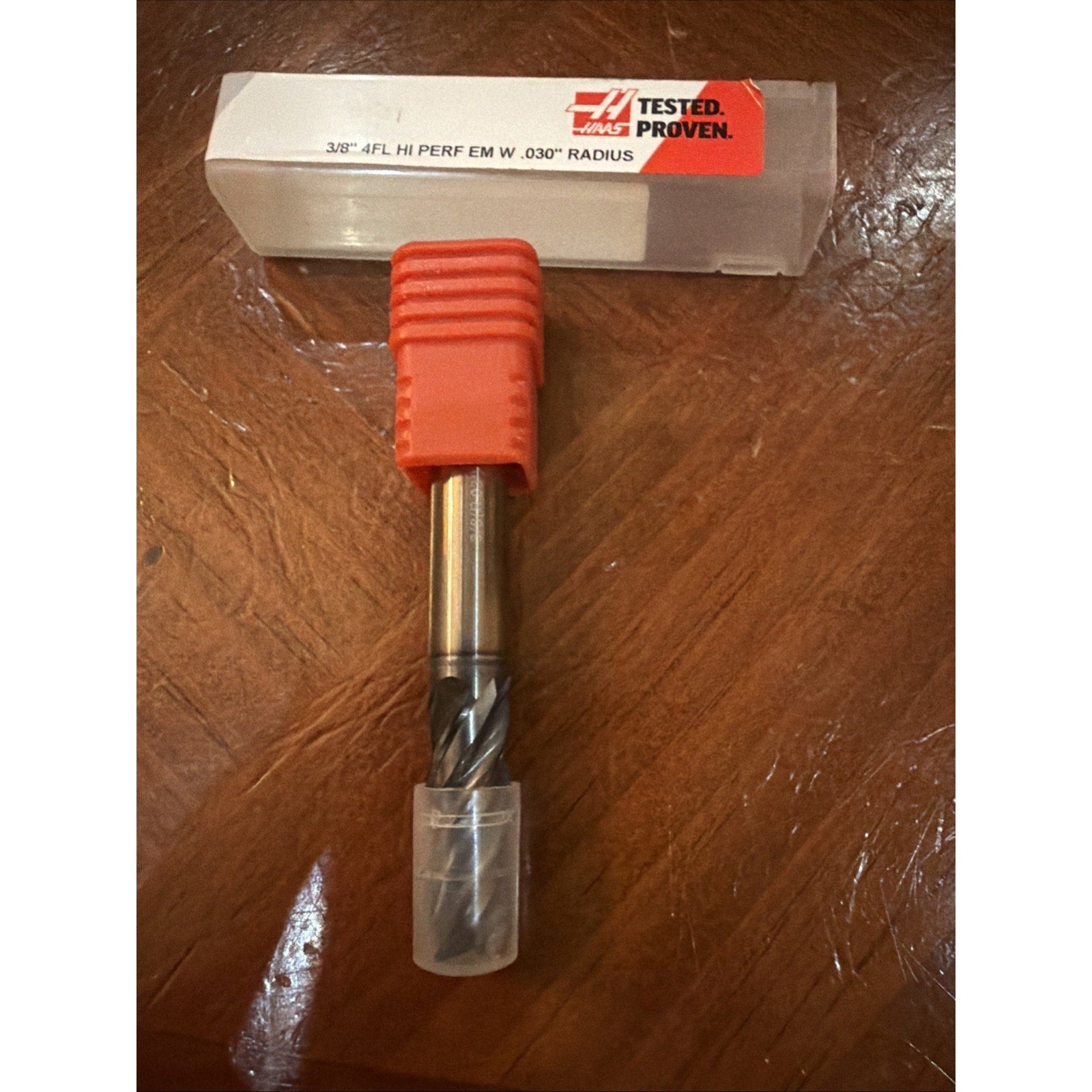 Metal cutting tool with spiral flutes, partially enclosed in clear and red plastic protective caps, displayed on a polished wooden surface alongside a labeled plastic storage case.