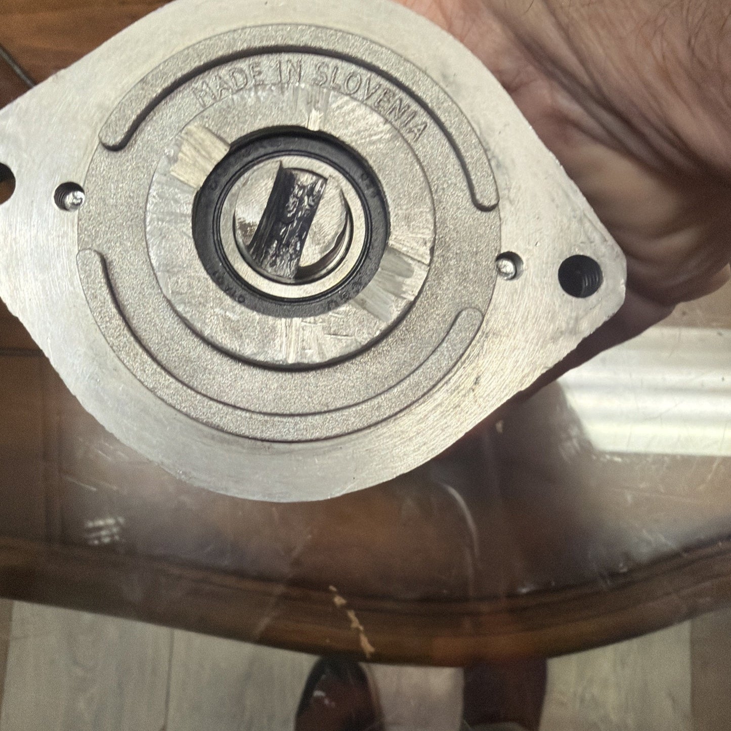 Close-up of a metallic circular mechanical component with a central shaft and engraved text "MADE IN SLOVENIA," held by a hand over a glass table.