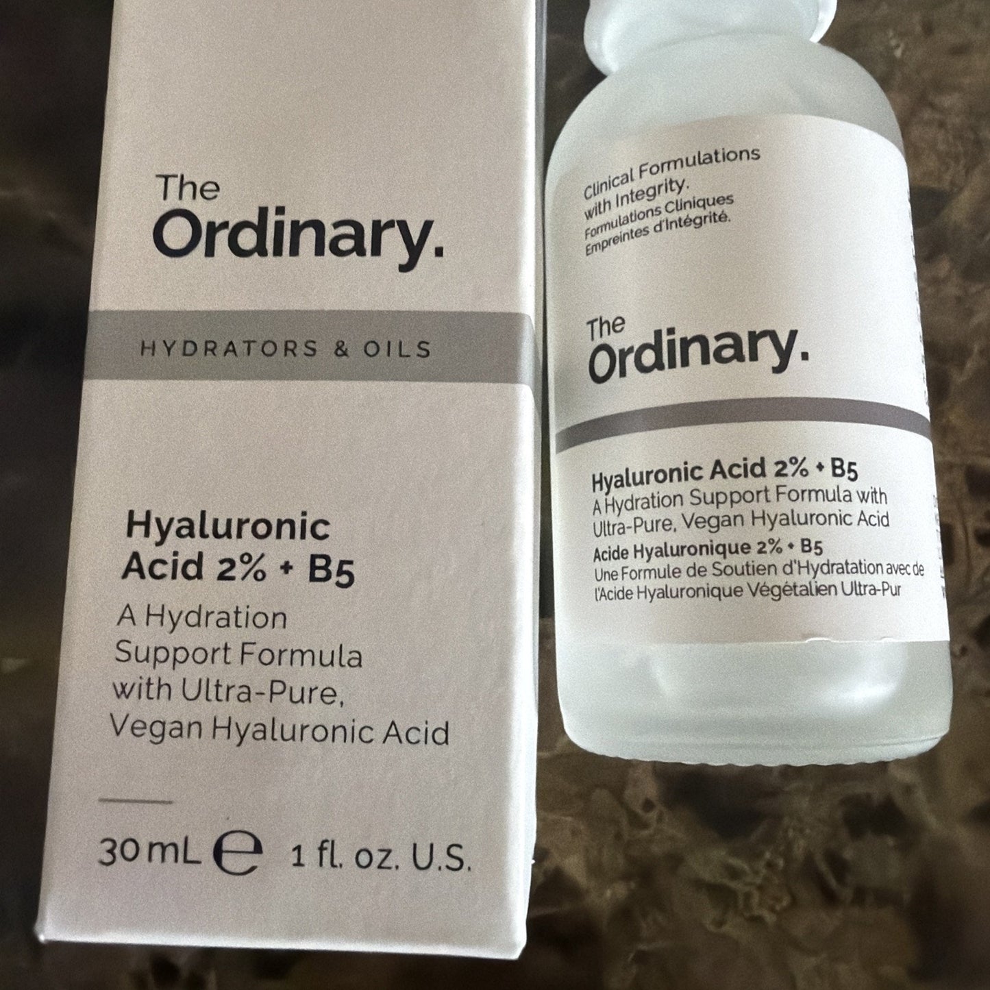 Frosted glass bottle and white box of The Ordinary Hyaluronic Acid 2% + B5 serum, featuring black text and a minimalist design, placed on a textured brown surface.