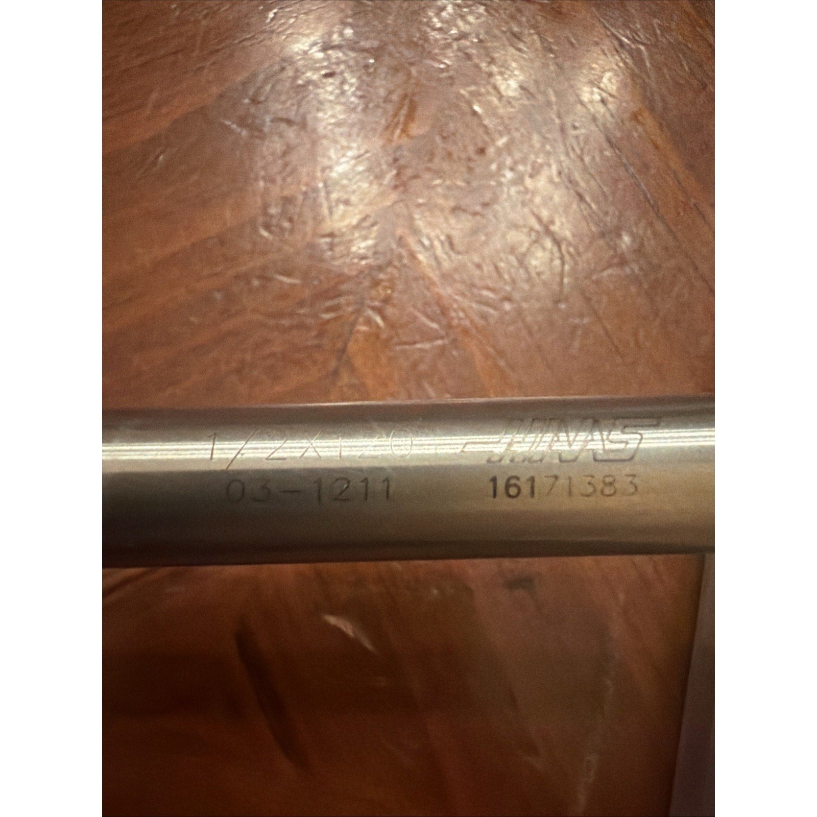 Close-up of a metal cylindrical tool with laser-engraved markings including the code "03-1211," a numbers sequence "16171383," and part of a brand name, resting on a brown wooden surface.