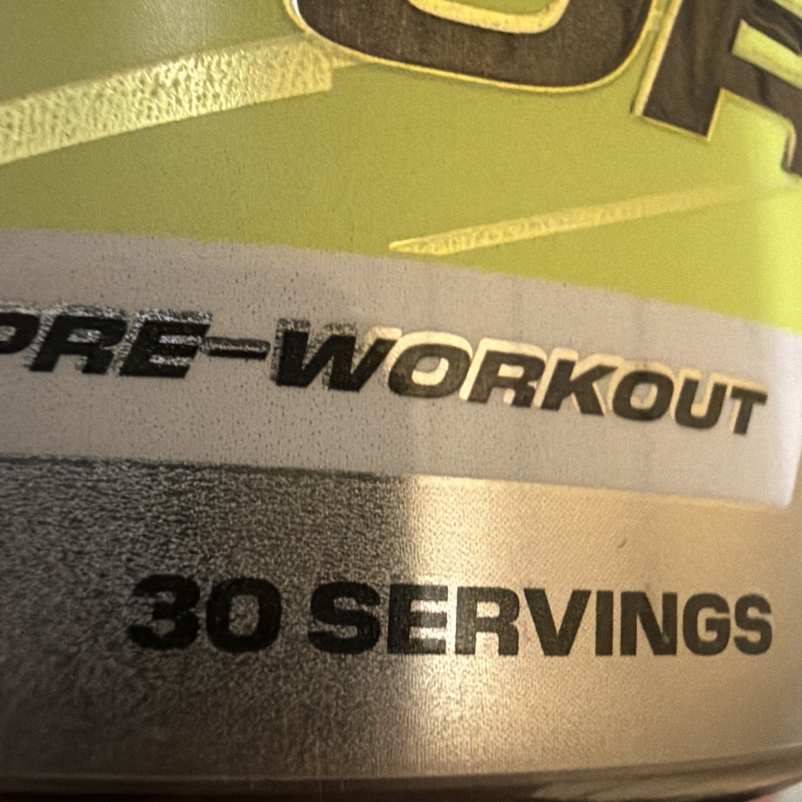 Close-up of a metallic container label displaying "PRE-WORKOUT" and "30 SERVINGS" in bold black text on a green and silver background.