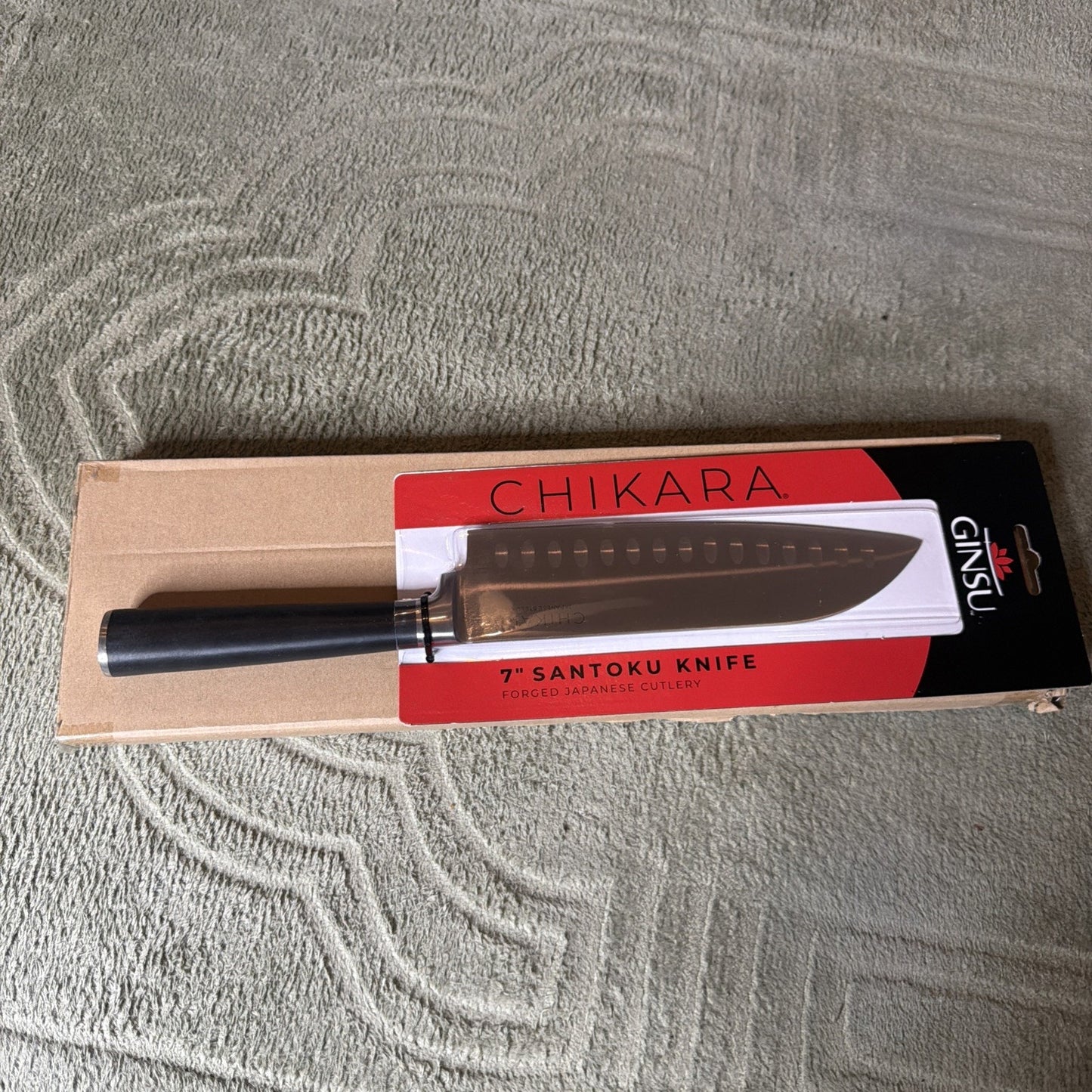 7-inch Santoku kitchen knife with a black handle displayed on red and black packaging labeled "Chikara" and "Ginsu," resting on a brown cardboard box on a textured surface.