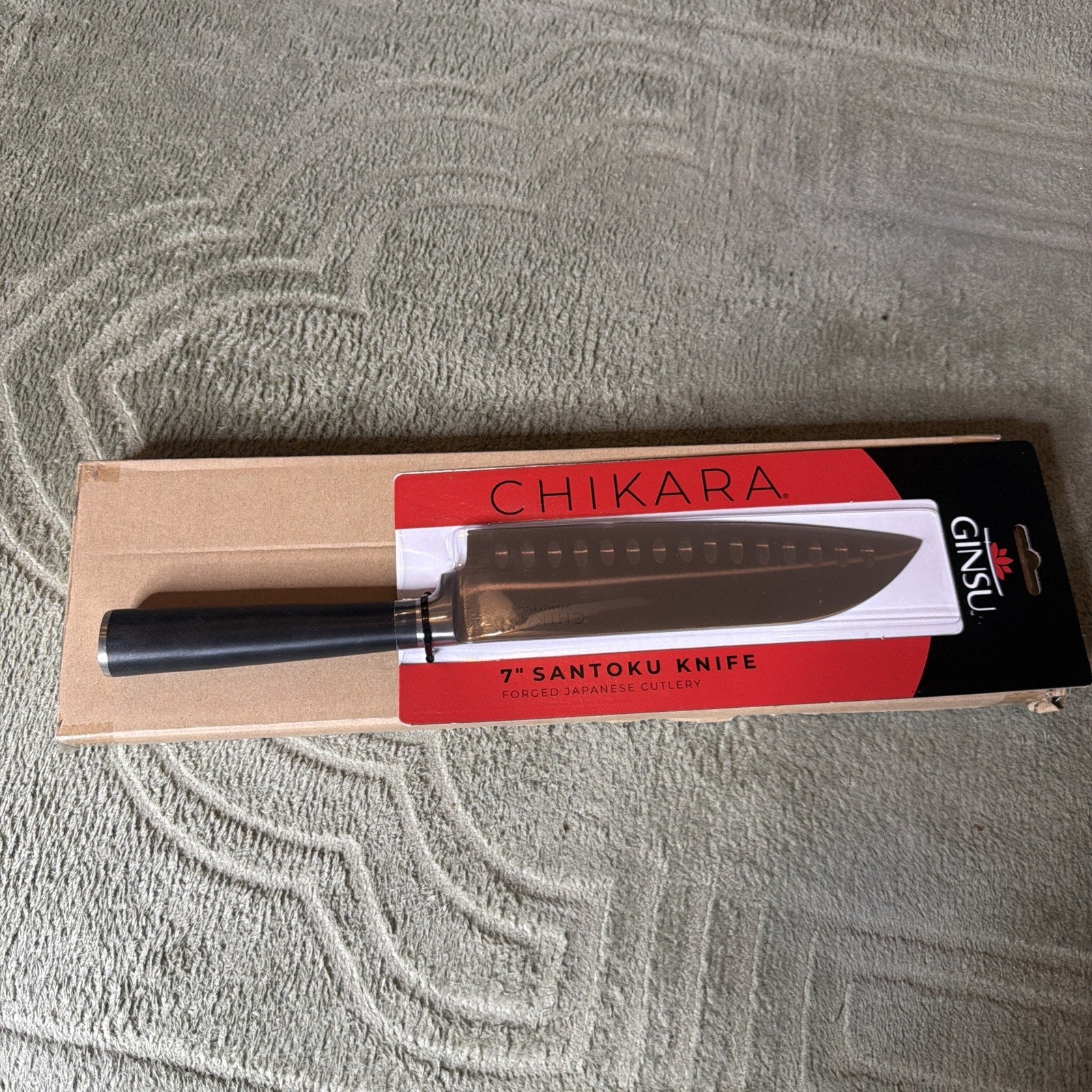 7-inch Santoku kitchen knife with a black handle displayed on red and black packaging labeled "Chikara" and "Ginsu," resting on a brown cardboard box on a textured surface.