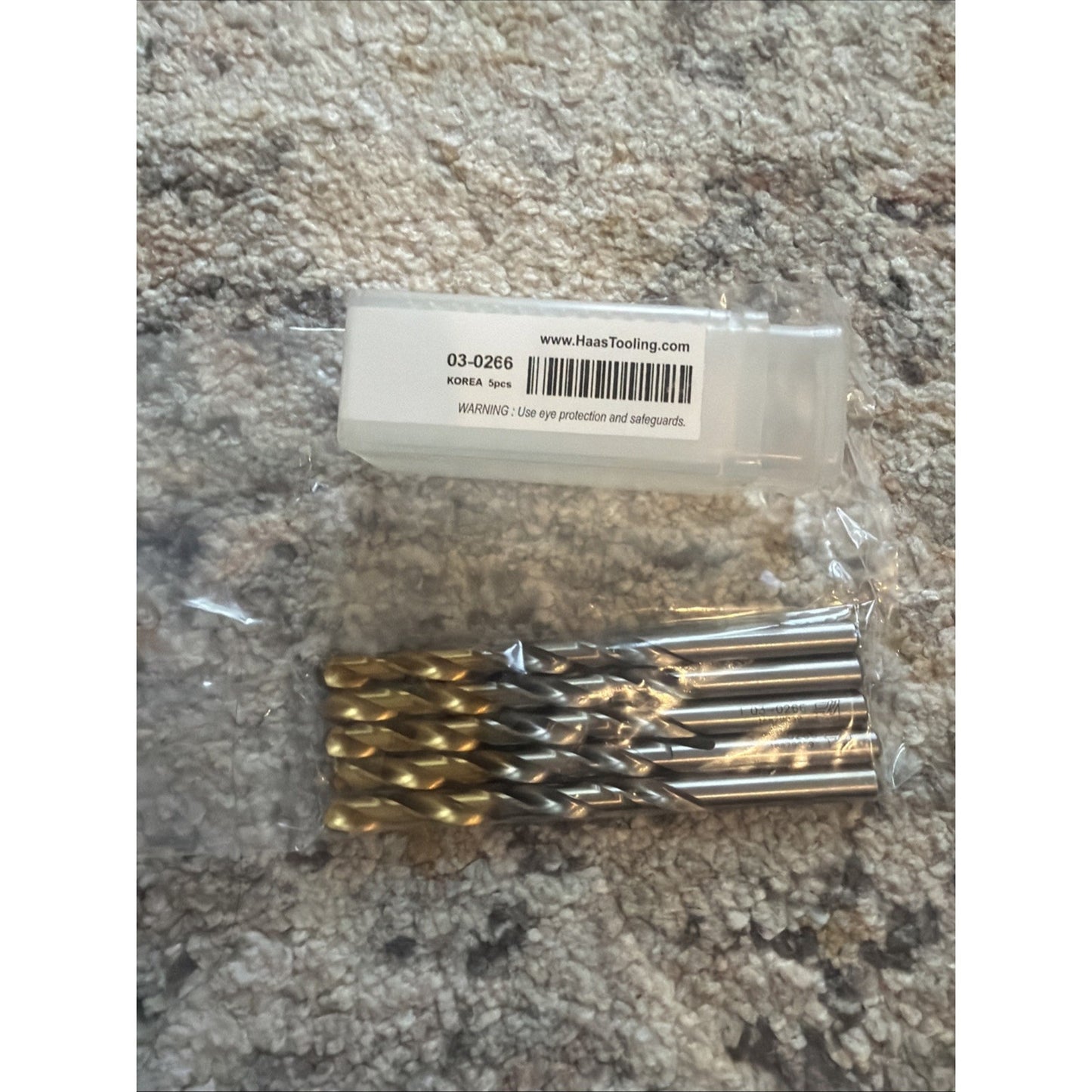 Set of eight gold-colored coated twist drill bits with silver shanks, wrapped in clear plastic, placed on a beige textured carpet next to a small labeled plastic case.