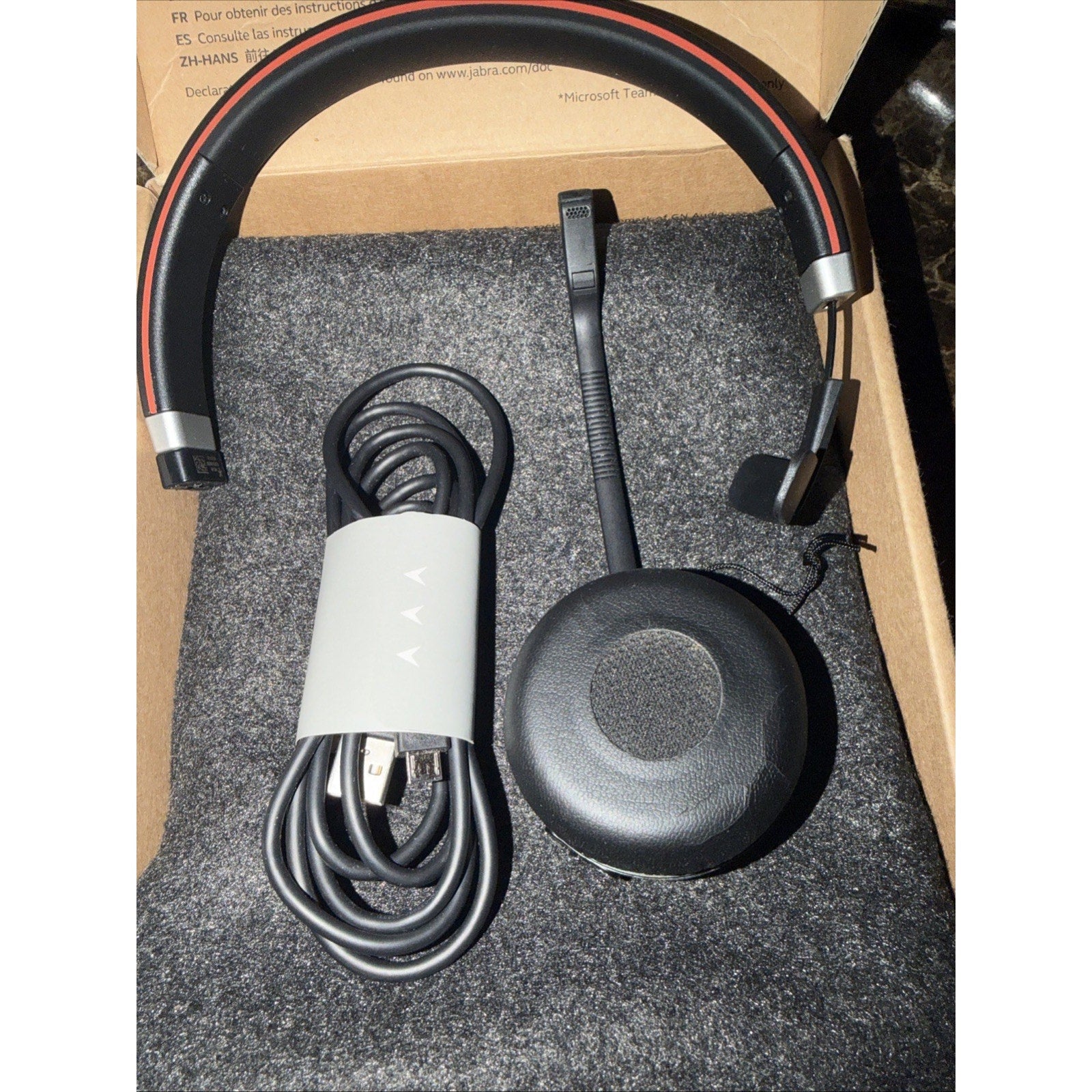 Black over-the-head style headset with a red trim, a flexible boom microphone, and a coiled USB charging cable, all placed inside a cardboard box with a soft black foam lining.