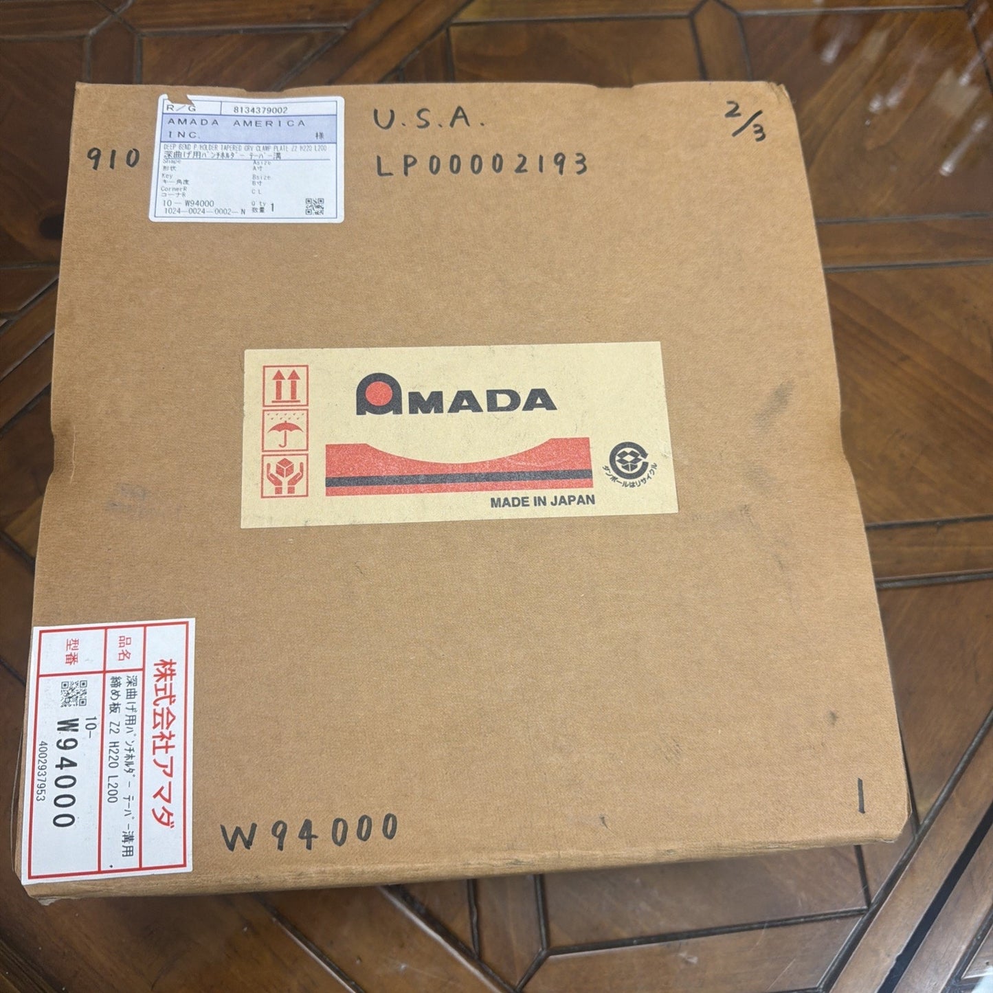 Brown cardboard shipping box with multiple labels, including a large AMADA logo labeled "Made in Japan," handwritten markings, and printed barcodes on a wooden floor.