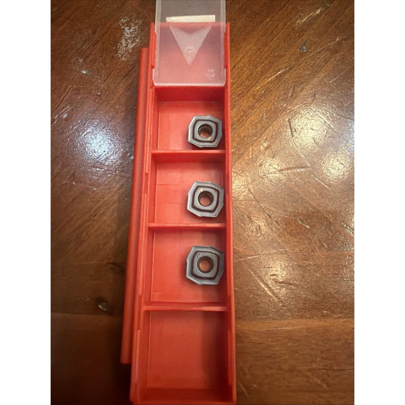 Red plastic storage container with three metal hexagonal inserts placed in separate compartments, set on a wooden surface with visible grain and lighting reflections.