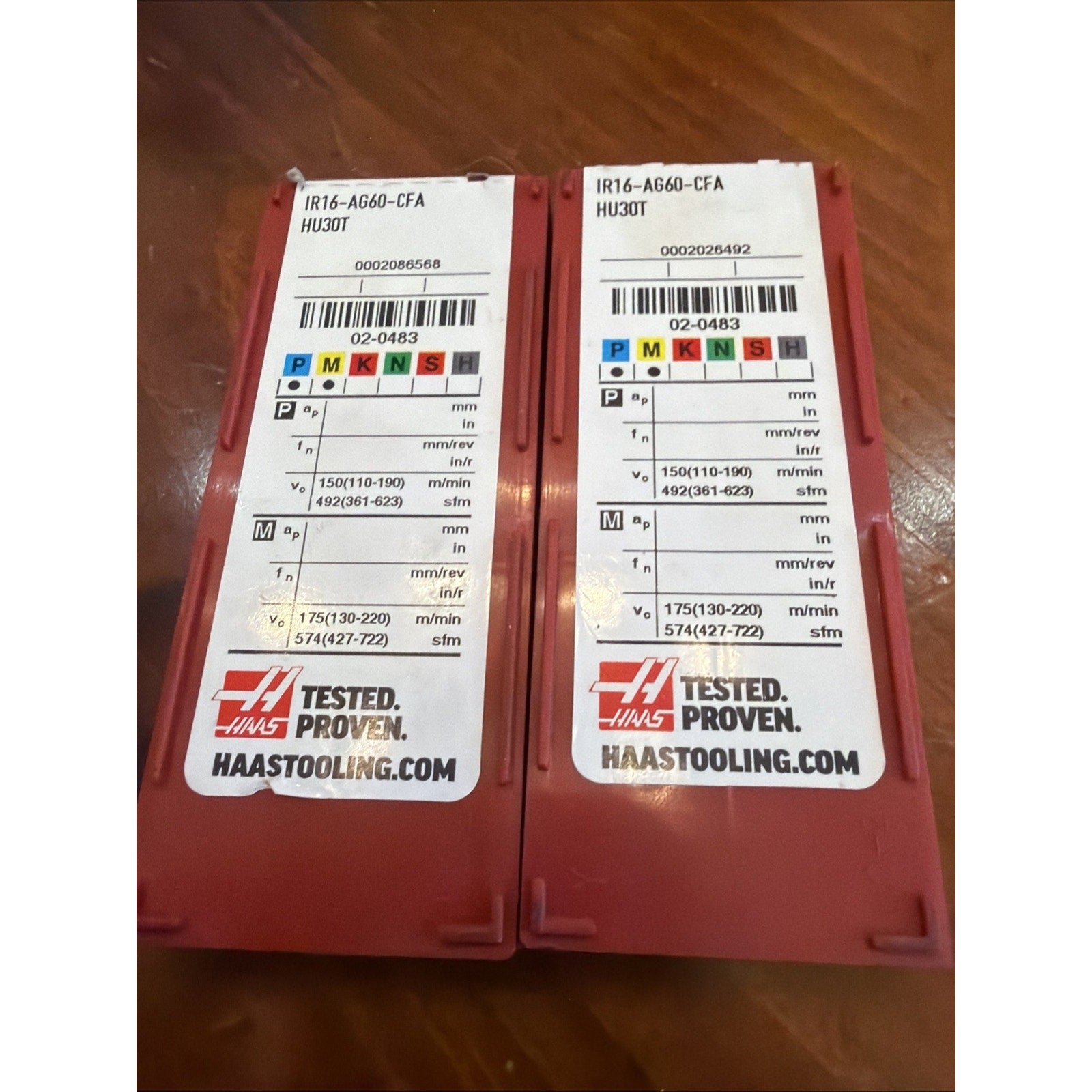 Two red plastic containers with white labels displaying technical specifications, colored coding for metal types, and Haas branding, placed side by side on a wooden surface.