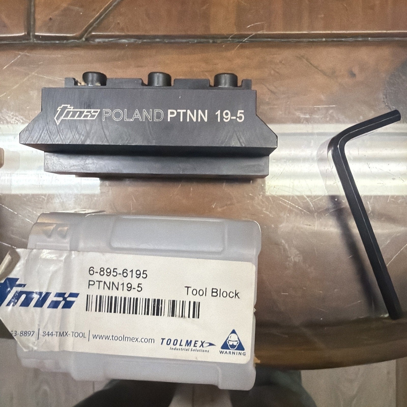 Dark metal tool block engraved with "TMG Poland PTNN 19-5," placed above a white label with product details, alongside a black L-shaped hex key on a reflective surface.