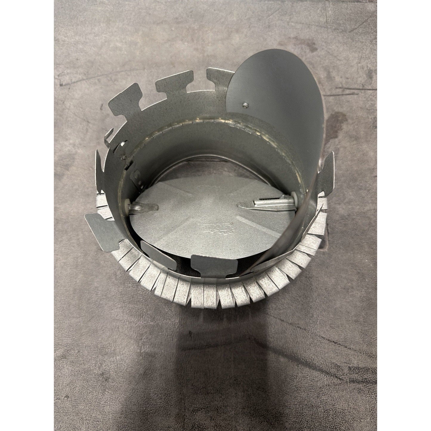 Metallic circular component with evenly spaced tabs around the outer edge and a hinged, semi-circular flap attached inside, placed on a gray surface.