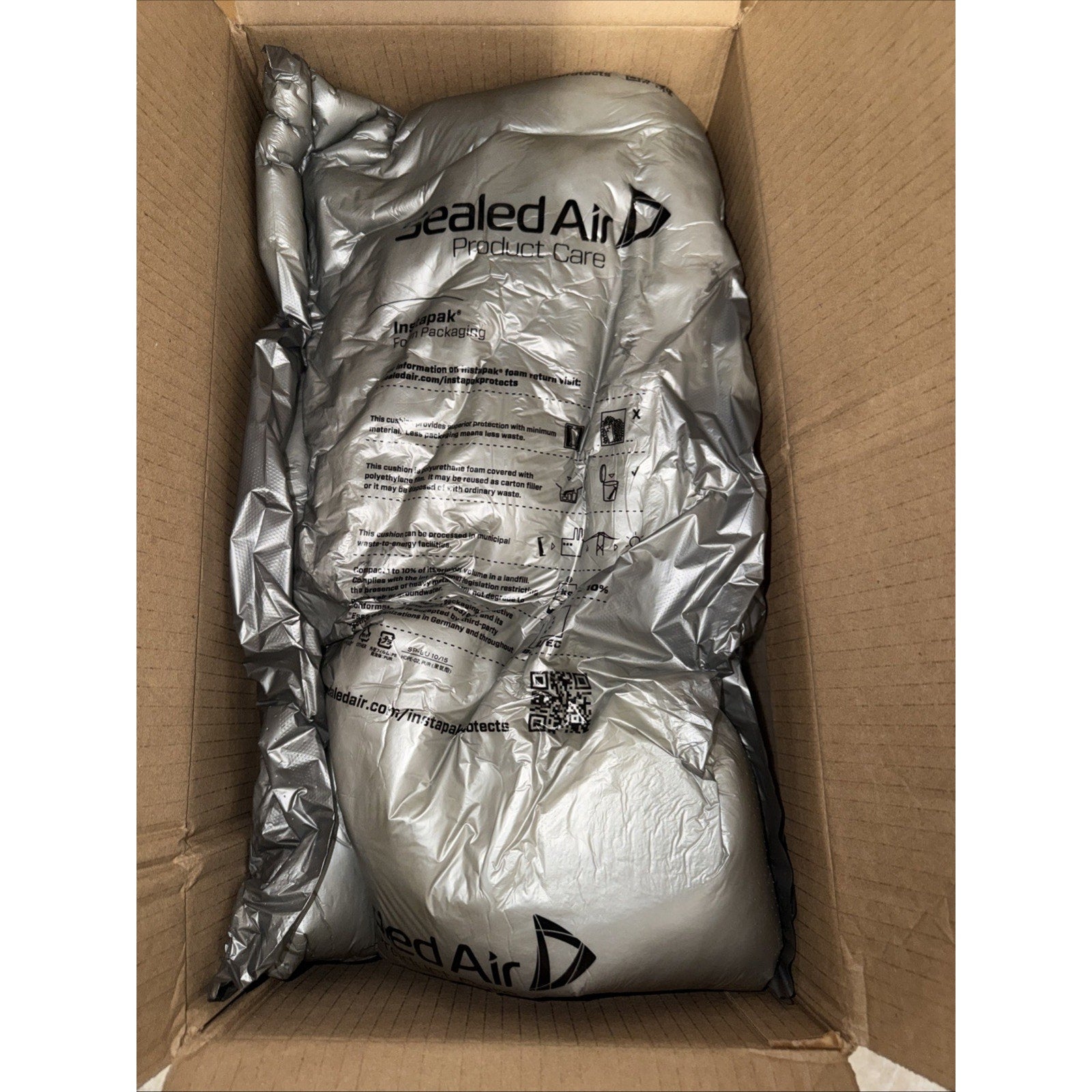 Inside a cardboard box, silver-colored inflatable air cushioning packaging material with printed instructions and QR code is used for protective padding.