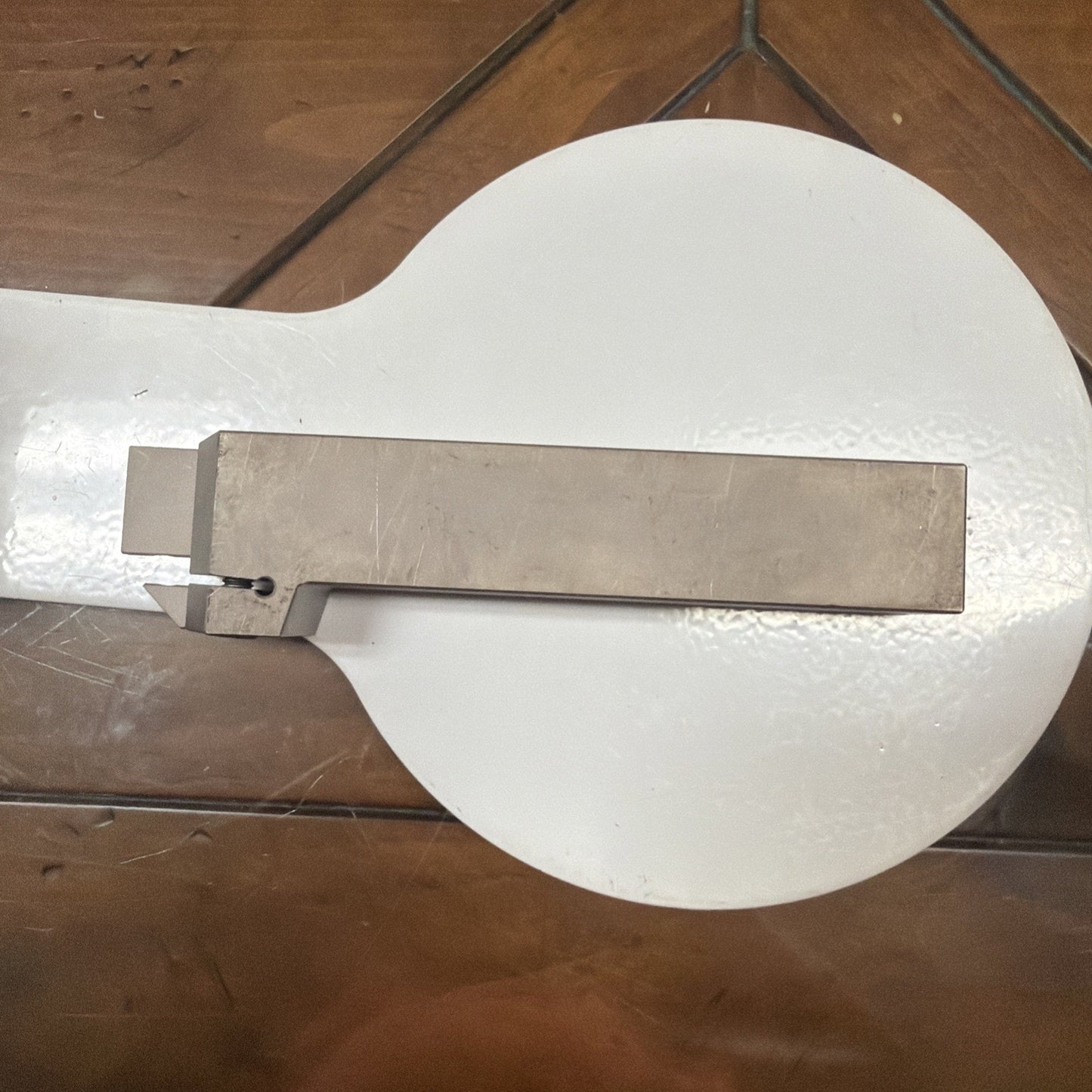 Rectangular metallic lathe cutting tool with a small square cutting edge and a hole near the tip, placed on a round white surface atop a wooden table.