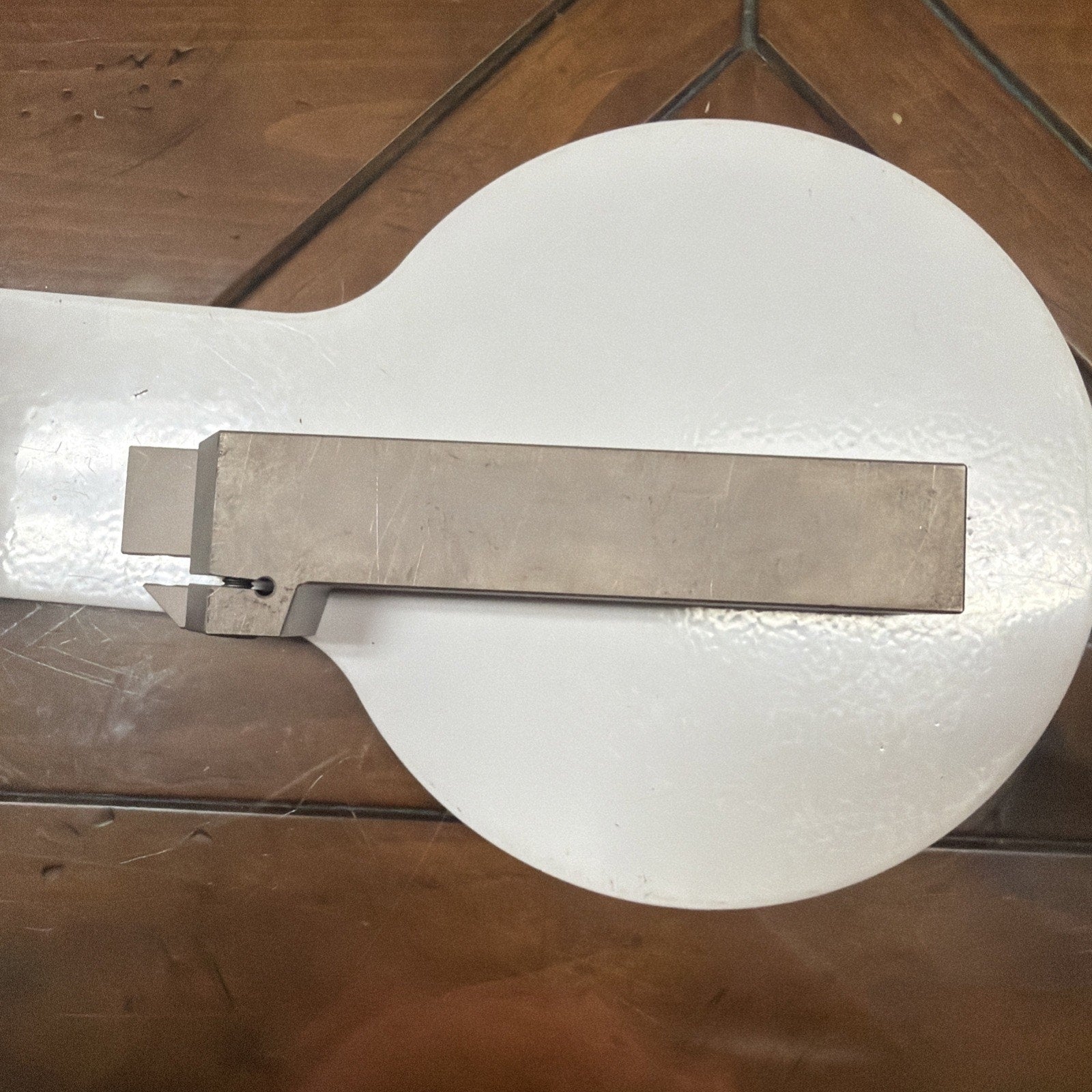 Rectangular metallic lathe cutting tool with a small square cutting edge and a hole near the tip, placed on a round white surface atop a wooden table.