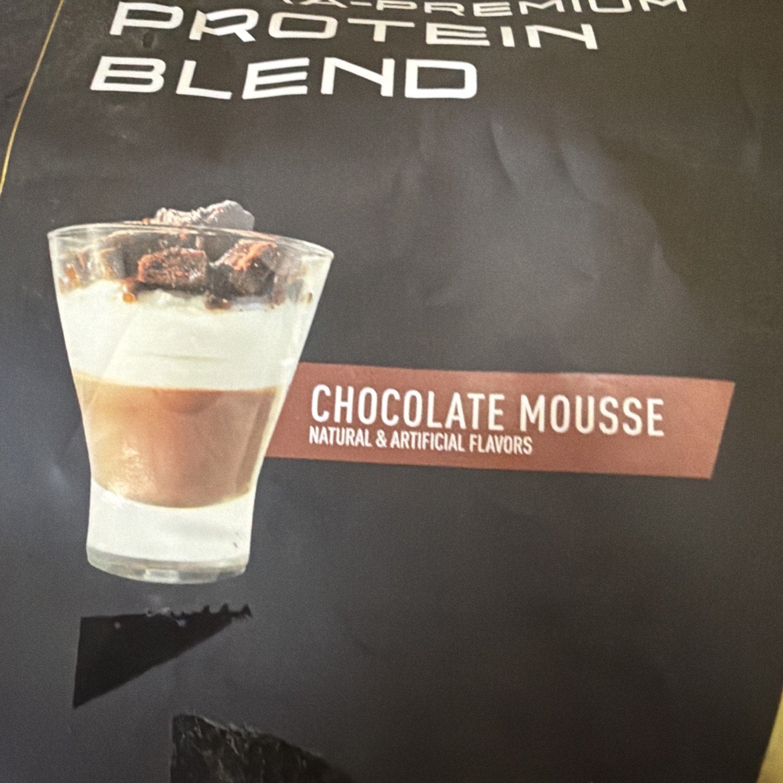 Clear glass filled with layered chocolate mousse dessert topped with chocolate chunks, shown on a dark background with "Chocolate Mousse" label and flavor description.