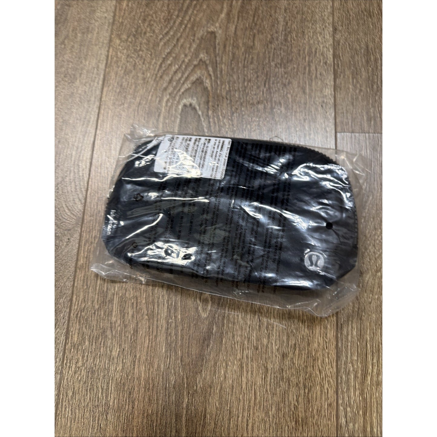 Small black pouch with a subtle logo on the bottom right corner, sealed in a clear plastic bag and placed on a wooden floor.