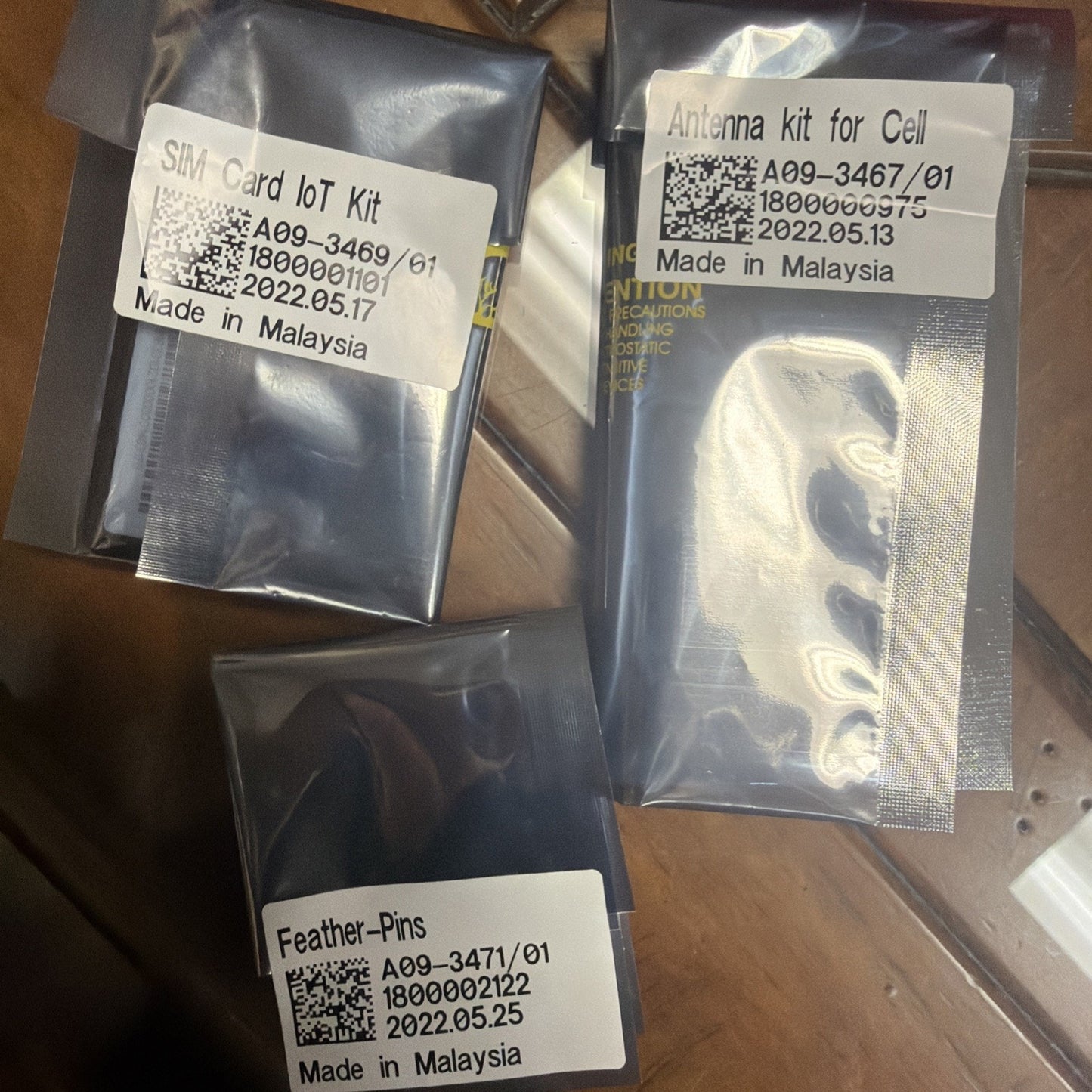 Three sealed plastic packages labeled "SIM Card IoT Kit," "Antenna kit for Cell," and "Feather-Pins," each with QR codes, product codes, and manufacturing dates, placed on a wooden surface.