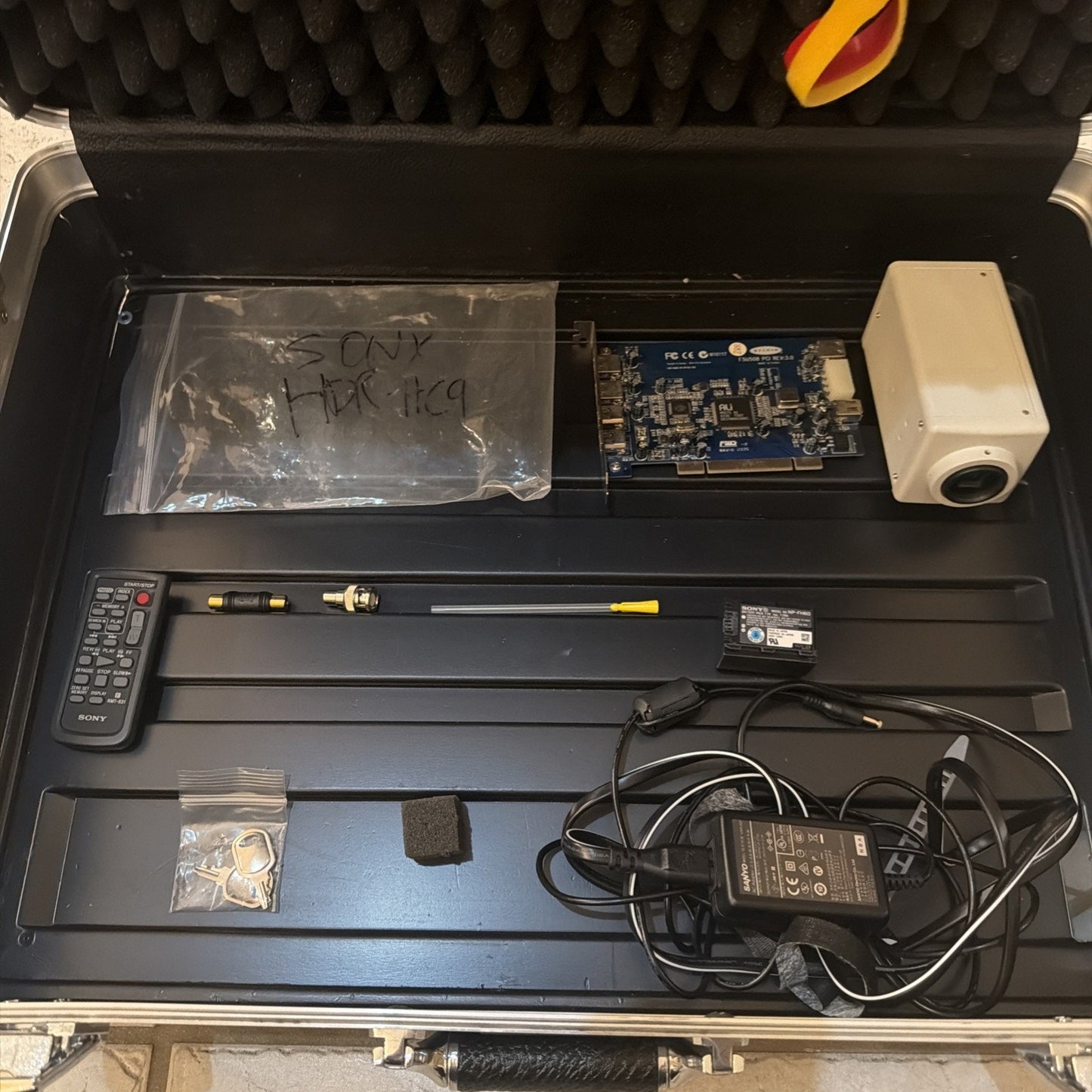 Open black equipment case containing an assortment of electronic components including a remote control, circuit board, white camera unit, power adapter with cables, small bag of metal keys, antenna rods, and a foam piece, all organized in custom foam compartments.