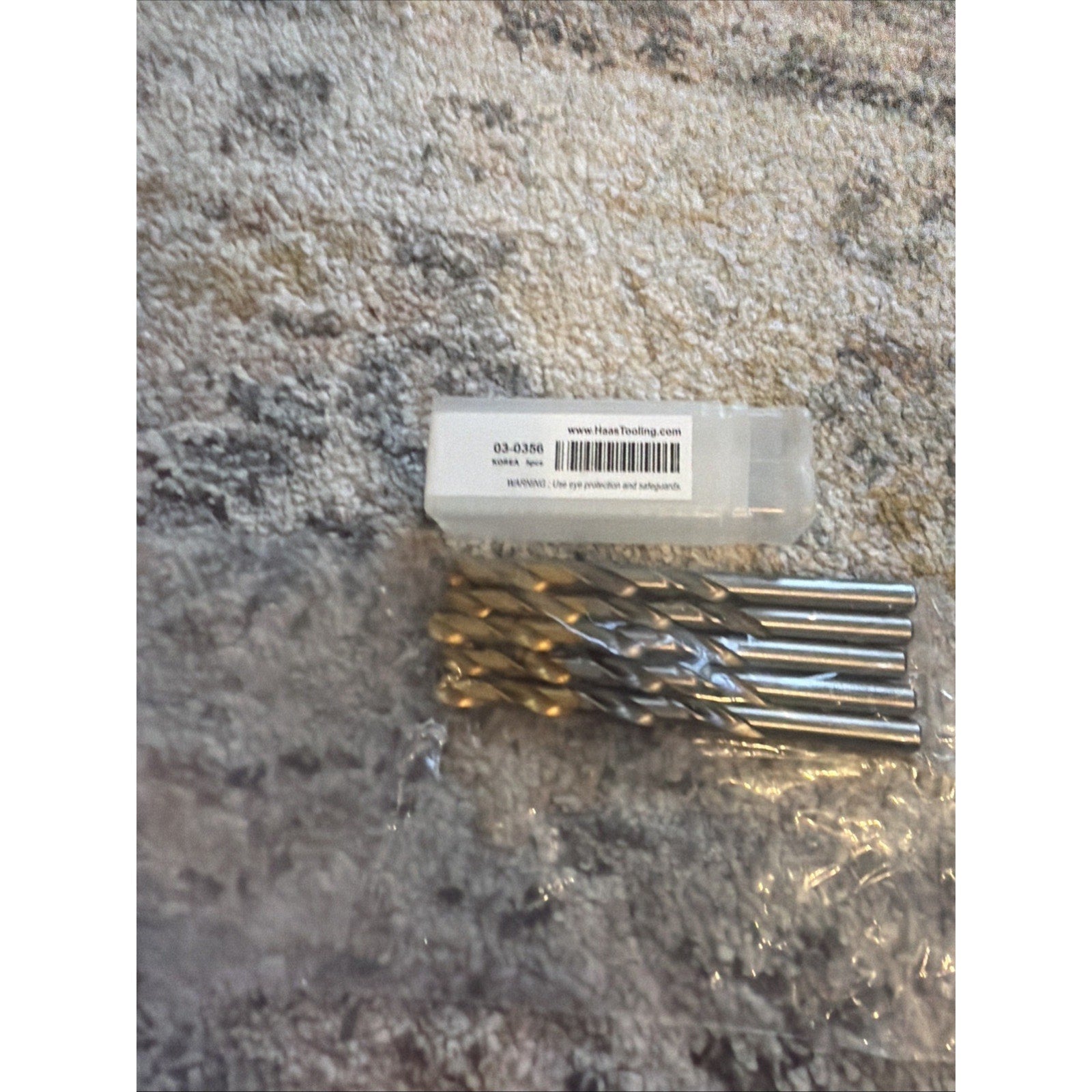 Set of five metal twist drill bits with spiral flutes, placed next to a small clear plastic box with a white label, all resting on a textured gray and beige surface.