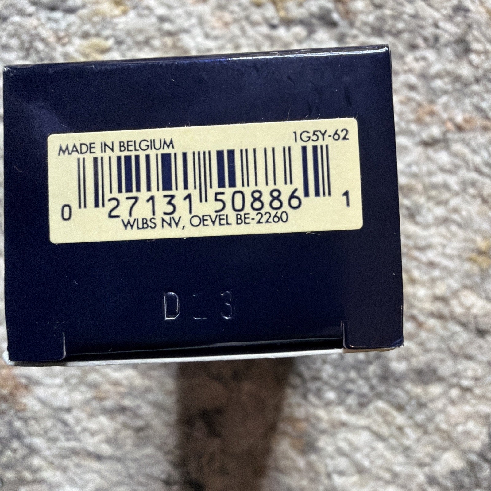 Close-up of the bottom of a dark blue box displaying a barcode label with the text "Made in Belgium," a product code, and manufacturer location information, stamped with "D-3.