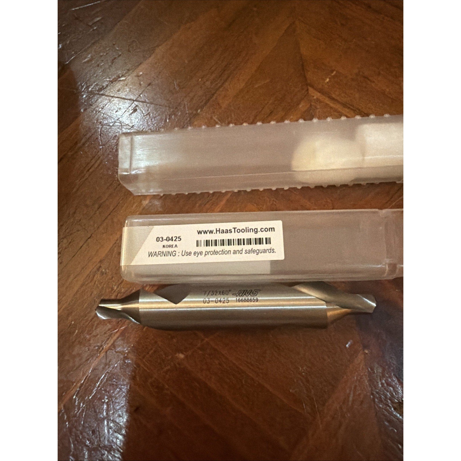 Double-ended metal cutting tool with tapered ends and engraved product code, resting on a wooden surface next to a clear plastic storage case with a warning label.