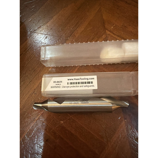 Double-ended metal cutting tool with tapered ends and engraved product code, resting on a wooden surface next to a clear plastic storage case with a warning label.