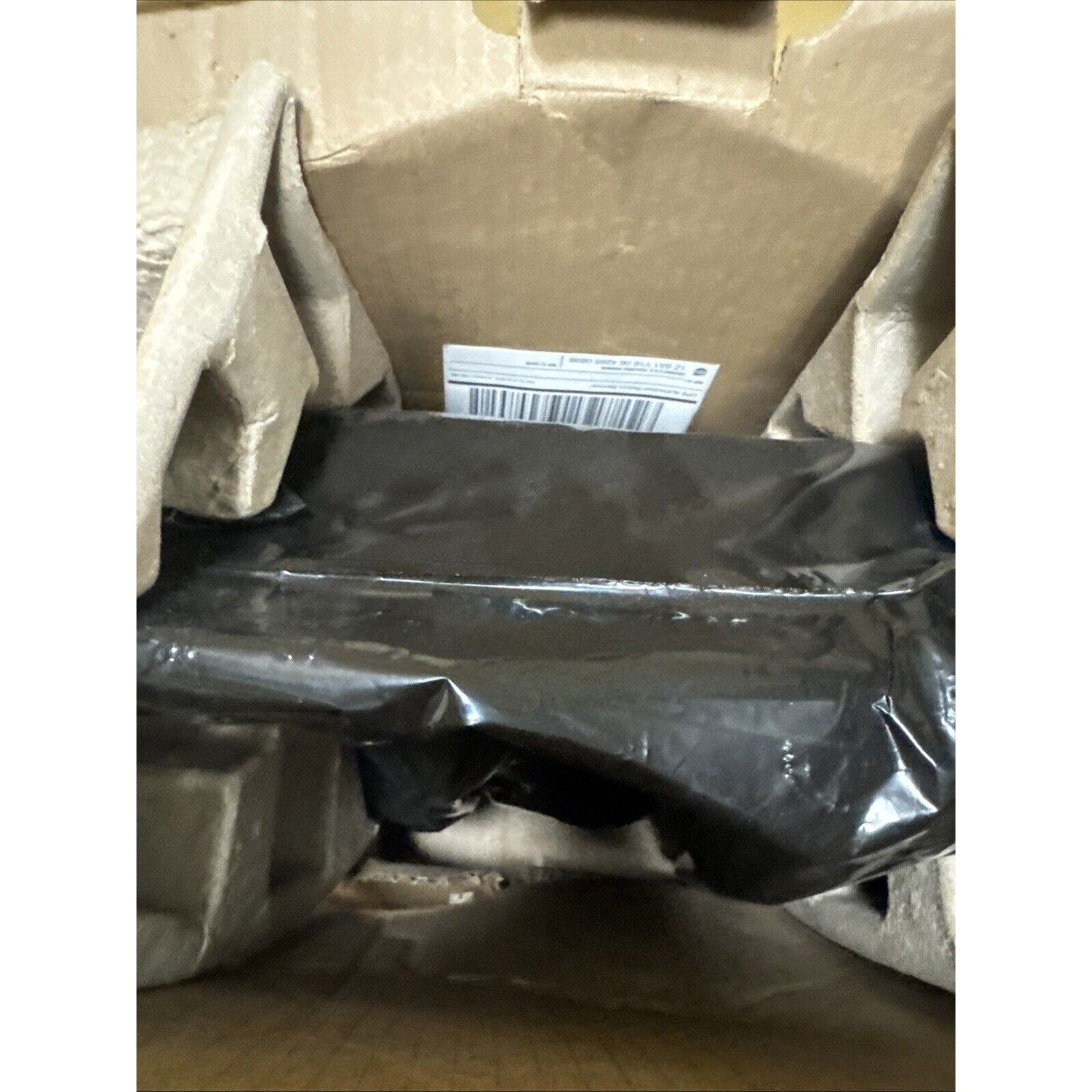 Brown cardboard box interior containing a black plastic-wrapped item secured by molded pulp protective inserts with a visible barcode label inside the box.