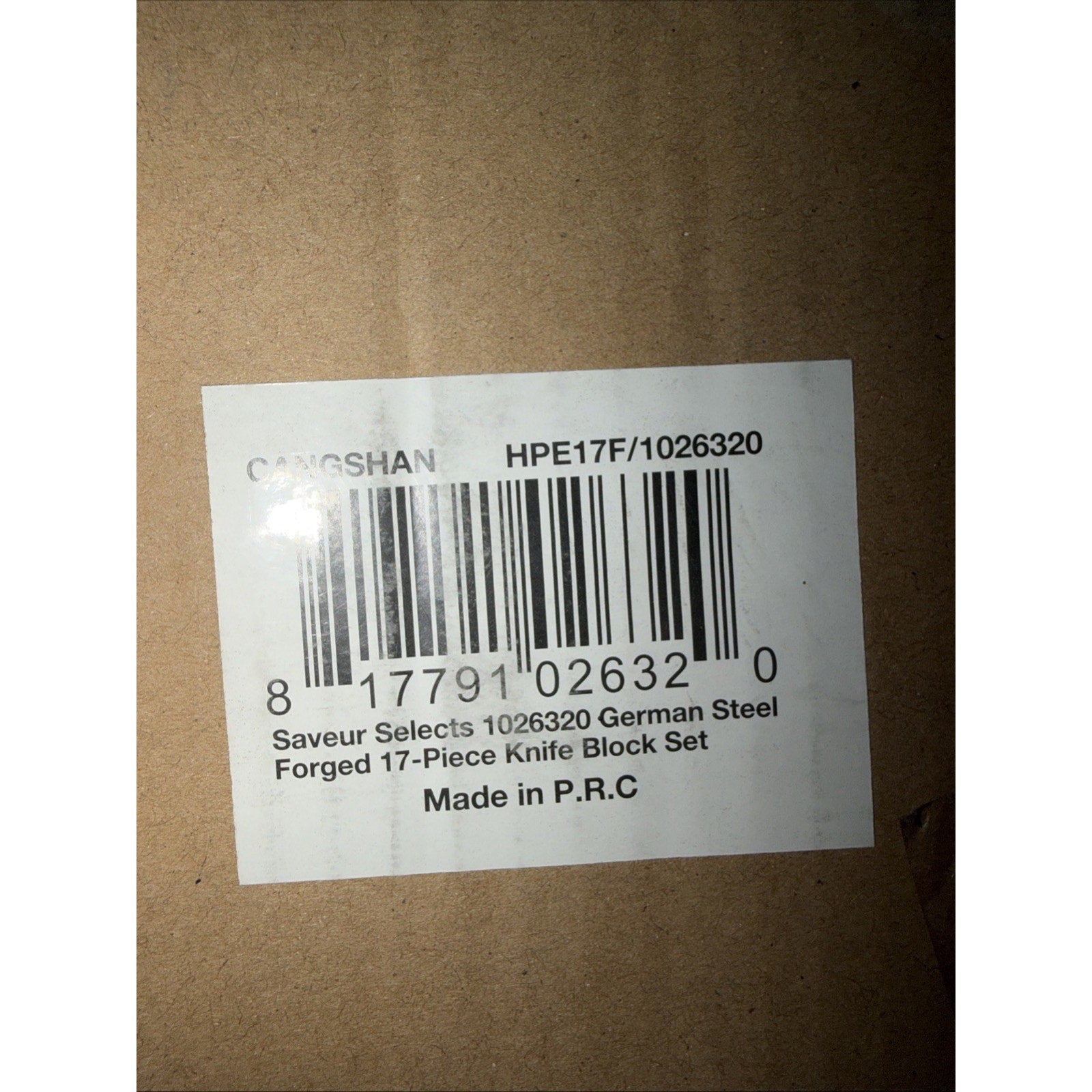 Close-up of a white product label on brown cardboard packaging showing a barcode and text listing a 17-piece German steel forged knife block set made in P.R.C.