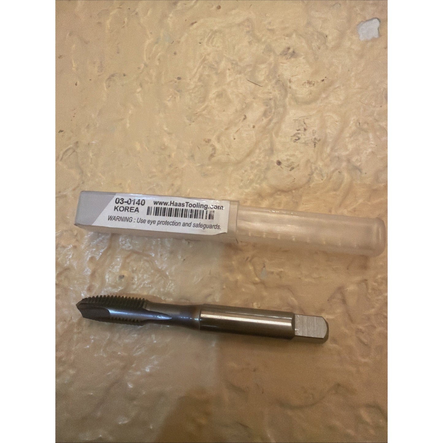 Single metal threading tap with sharp flutes and a square drive end, placed on a textured beige surface next to a transparent plastic storage tube labeled with product details and safety warning.