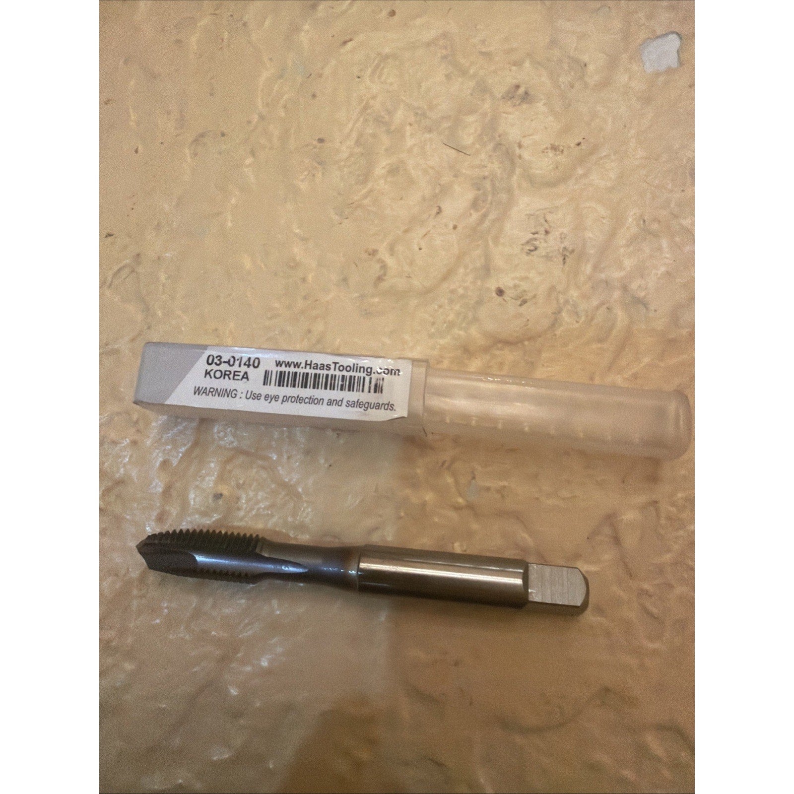 Single metal threading tap with sharp flutes and a square drive end, placed on a textured beige surface next to a transparent plastic storage tube labeled with product details and safety warning.