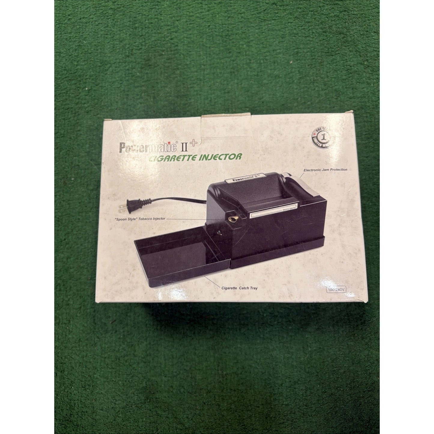 Box packaging of a black electric cigarette injector machine with a built-in cigarette catch tray, displayed on a green textured surface.