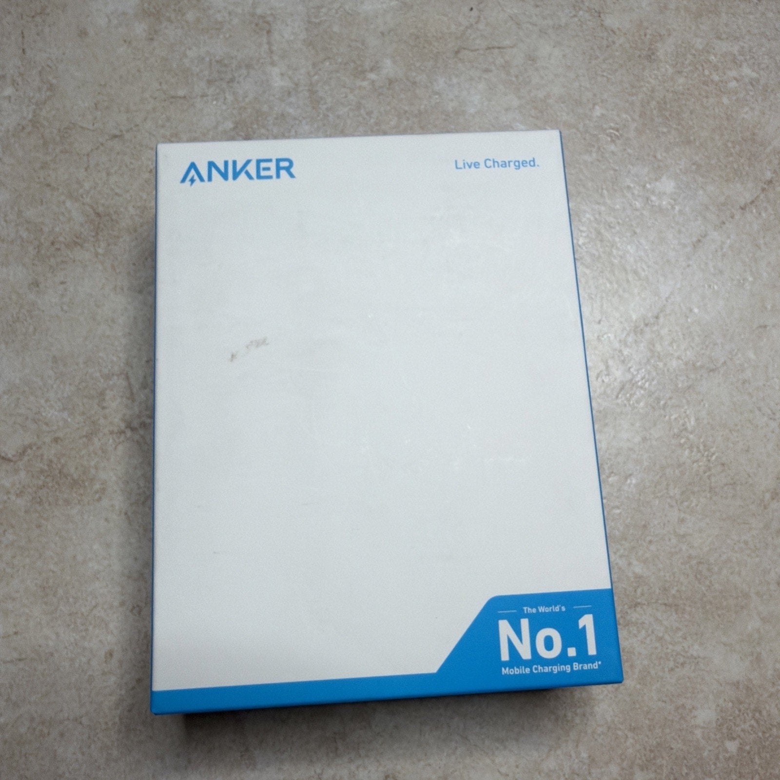 White Anker product box with blue accents, featuring the brand name in the top left corner and a blue banner in the bottom right highlighting it as the world's number one mobile charging brand, placed on a beige textured surface.