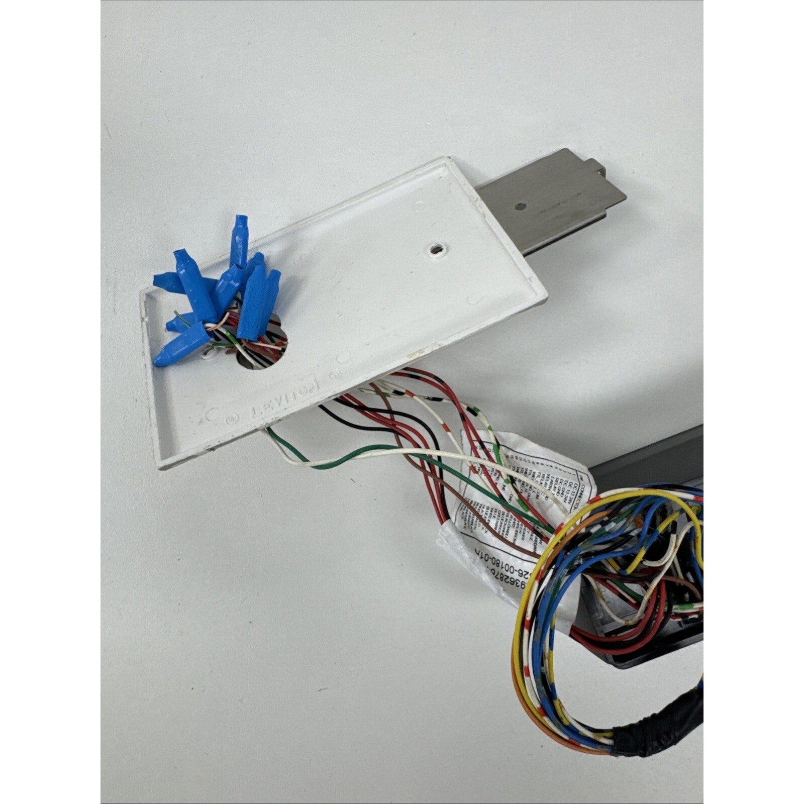 White rectangular plastic panel with multiple blue wire connectors attached to exposed multi-colored electrical wires, connected to a gray metal bracket on a white surface.