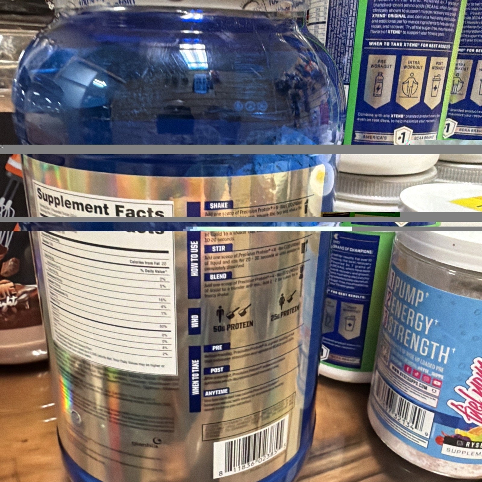 Back view of a large blue container with a metallic label displaying supplement facts, usage instructions for shake, stir, and blend, and protein content details, surrounded by other nutrition and supplement containers on a wooden surface.