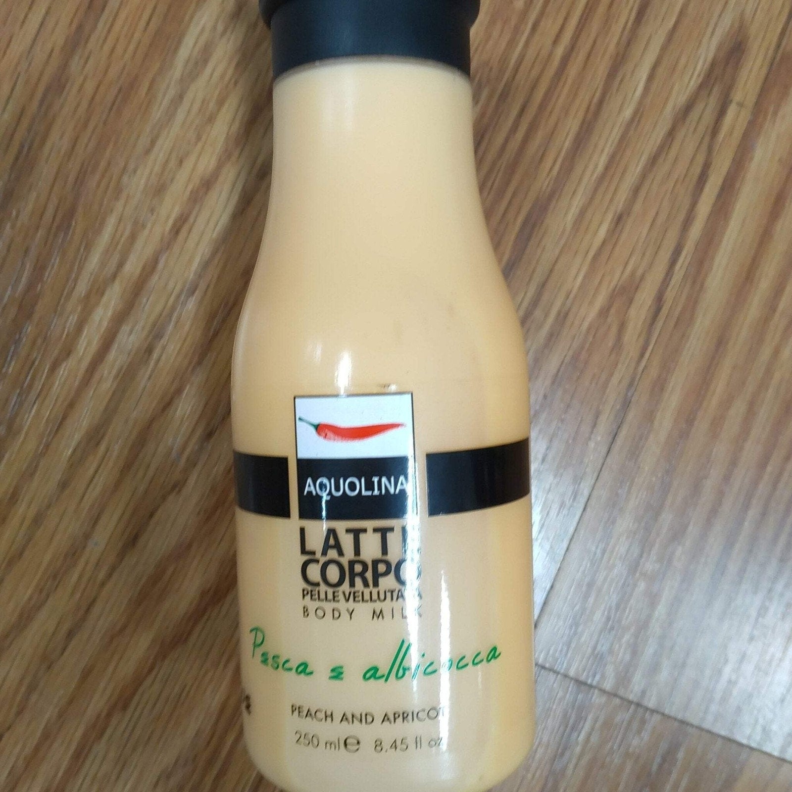 Cream-colored plastic bottle of Aquolina body milk with black cap, labeled in Italian and English indicating peach and apricot scent, placed on a wooden surface.