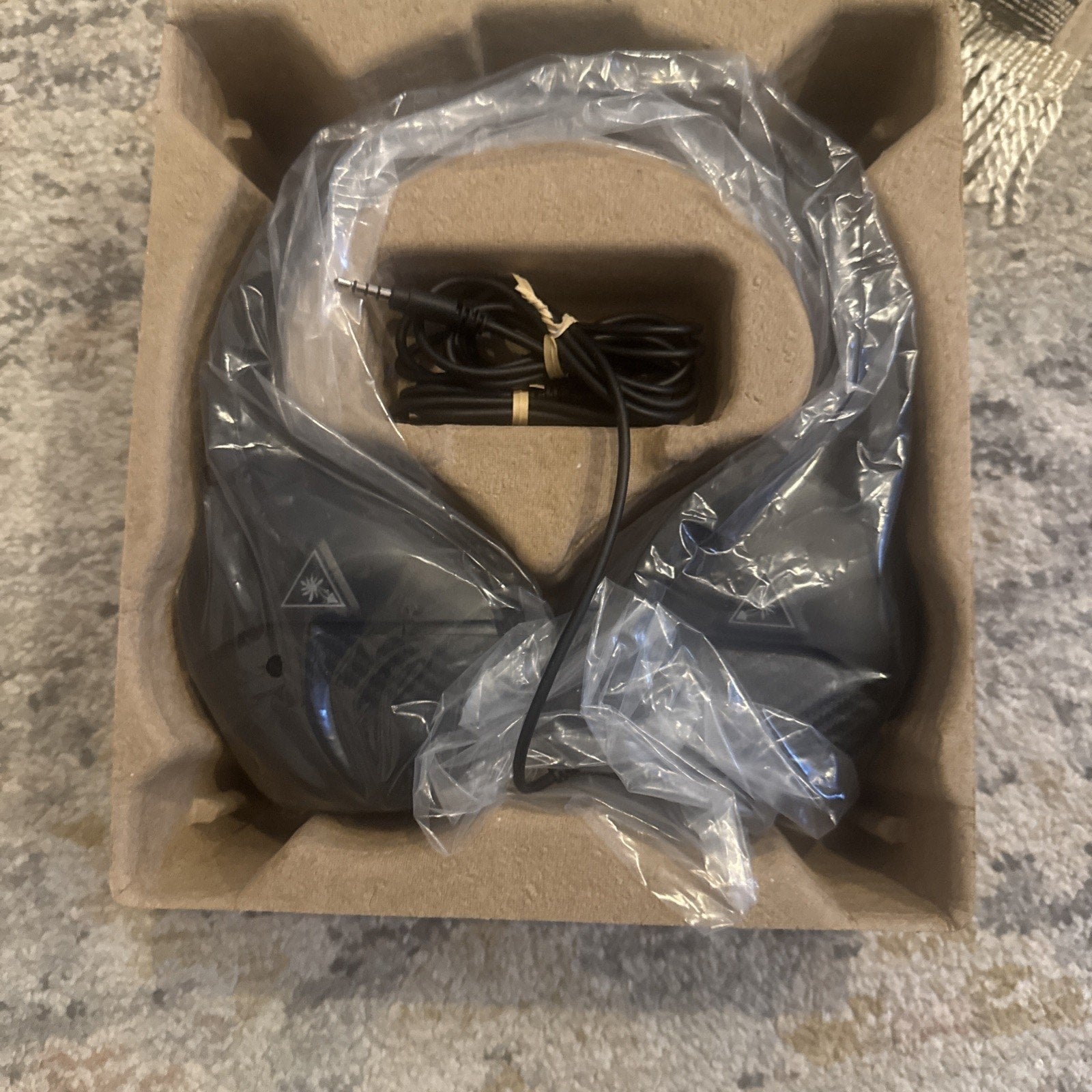Black over-ear headphones wrapped in plastic inside brown molded cardboard packaging, with a coiled black audio cable secured by a twist tie.