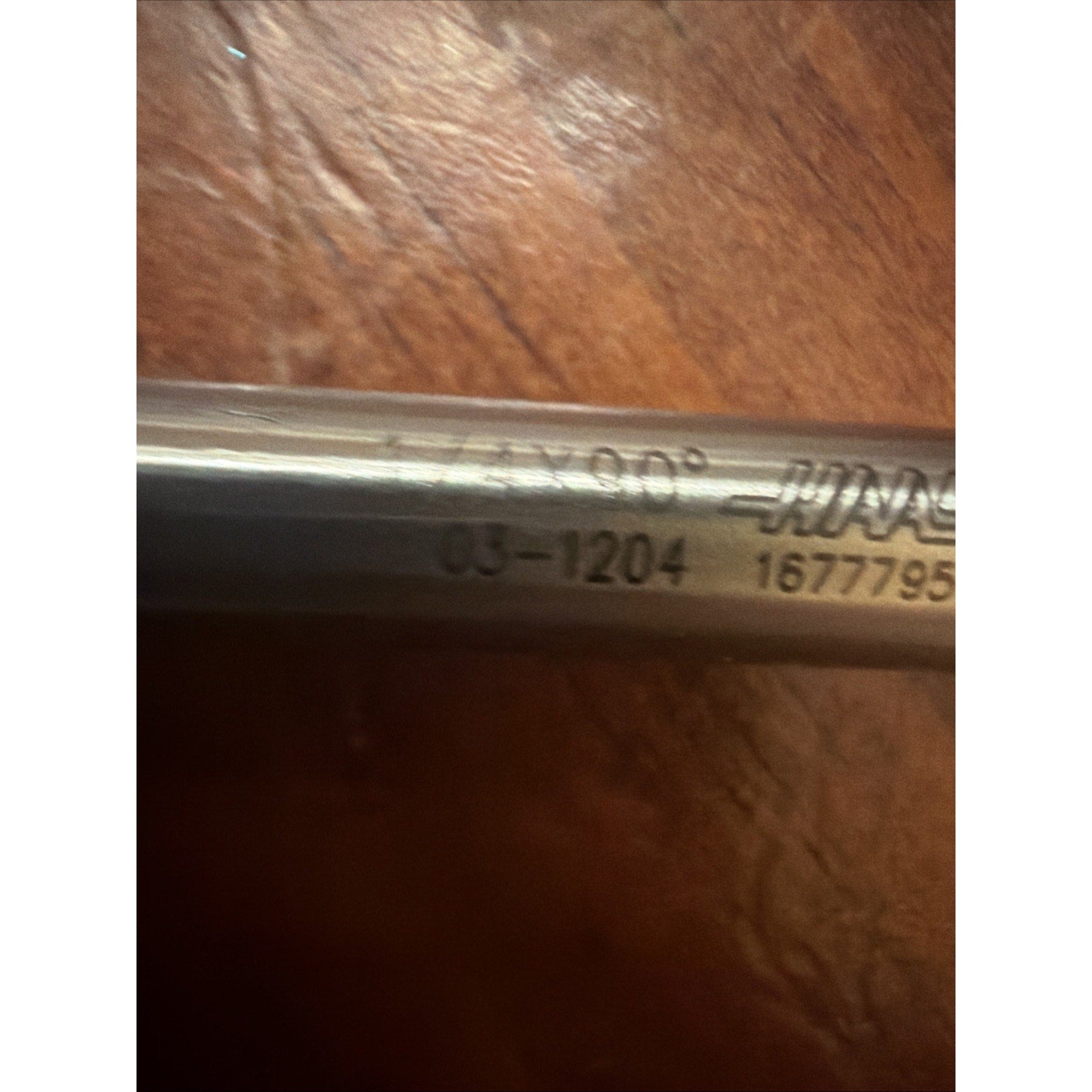 Close-up of a metal tool shaft engraved with markings including "03-1204" and a serial number, placed on a textured wooden surface.