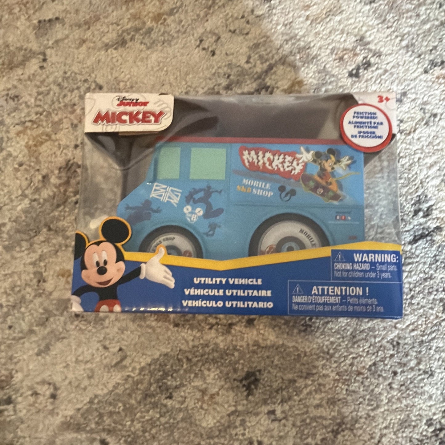 Blue toy utility vehicle with Mickey Mouse and Goofy graphics, packaged in a clear box with Disney Junior Mickey branding and safety warnings on a carpeted surface.