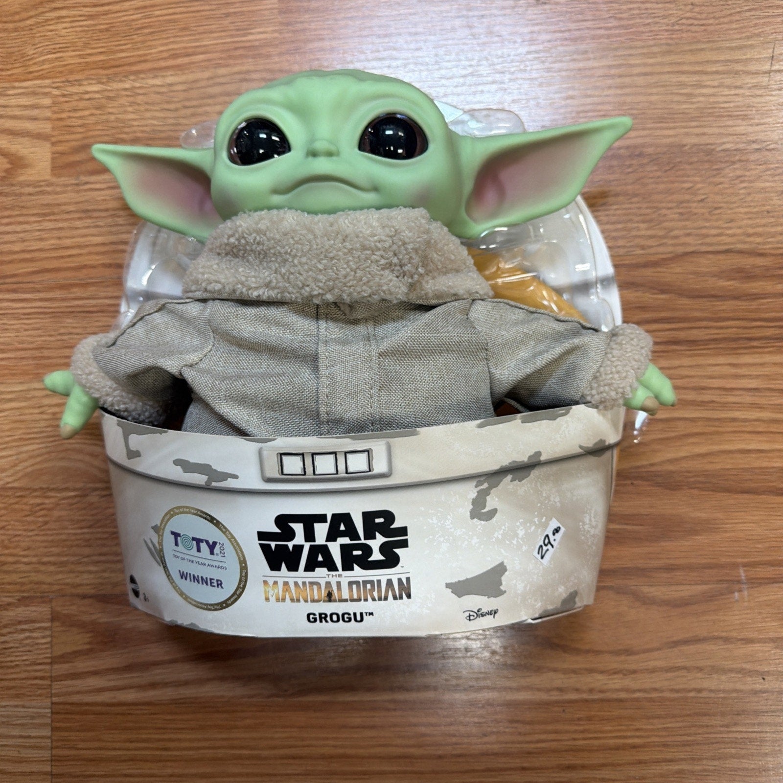 Plastic and fabric Grogu figure from Star Wars The Mandalorian, dressed in a beige coat with fleece collar, displayed in branded packaging on a wooden surface.