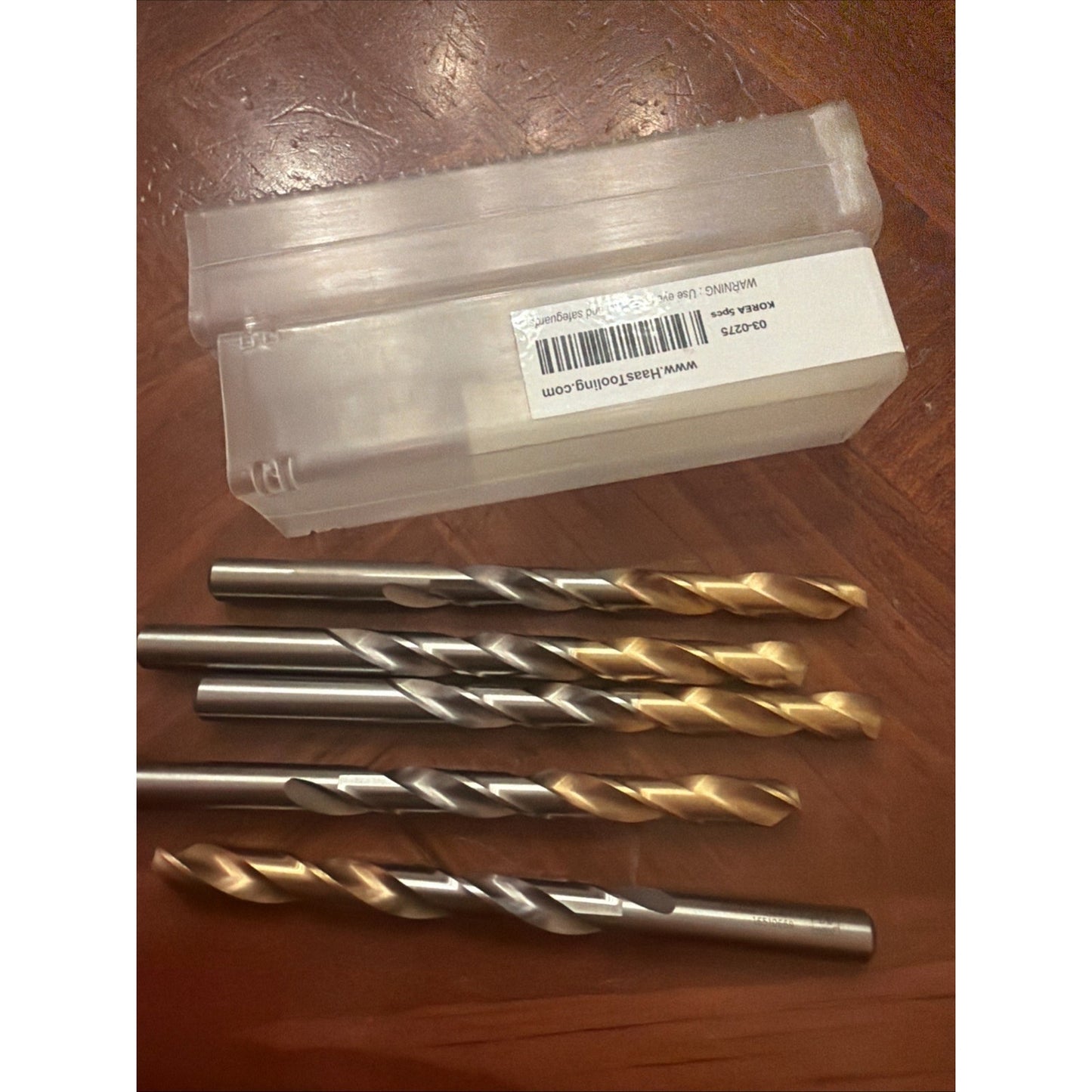 Five drill bits with a mix of silver and gold-colored tips arranged on a wooden surface next to a translucent plastic case with a white label and barcode.