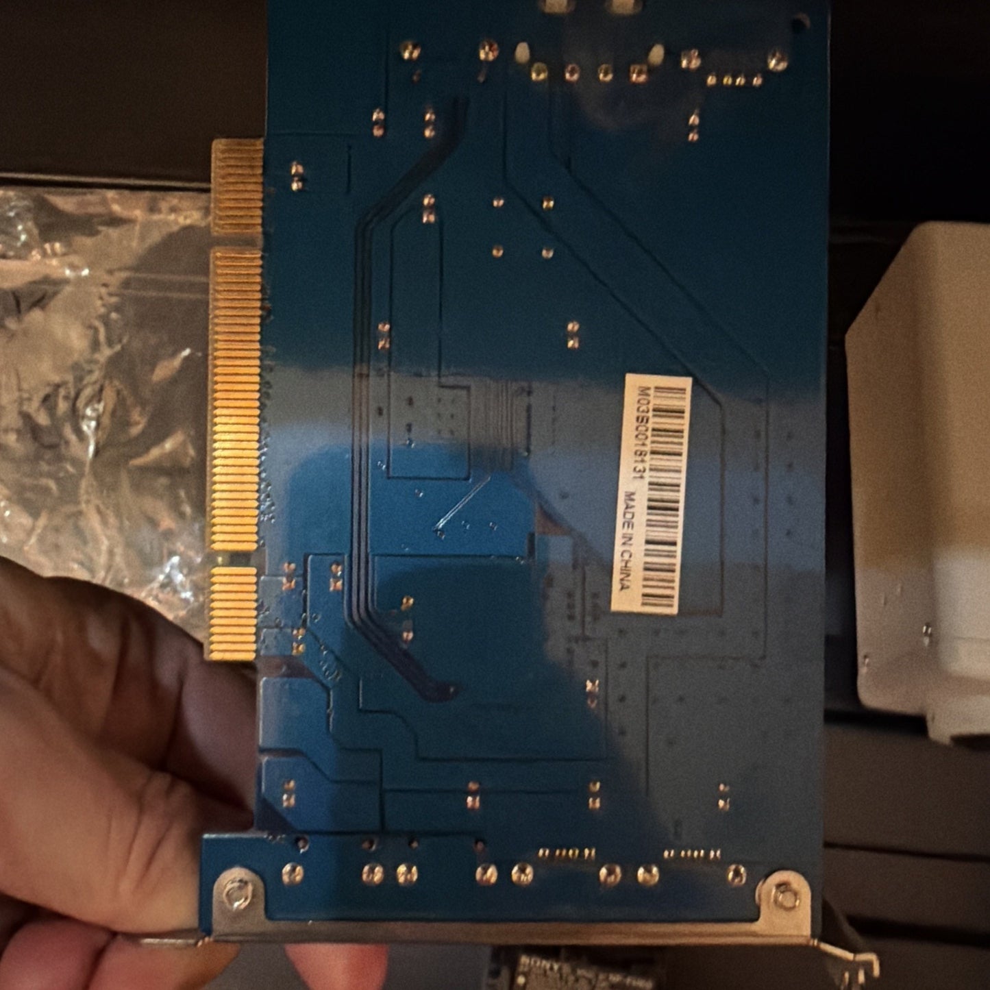 Close-up of a blue circuit board with a gold edge connector, small solder points, and a white barcode label with "Made in China" text, held by a hand against a dark background.