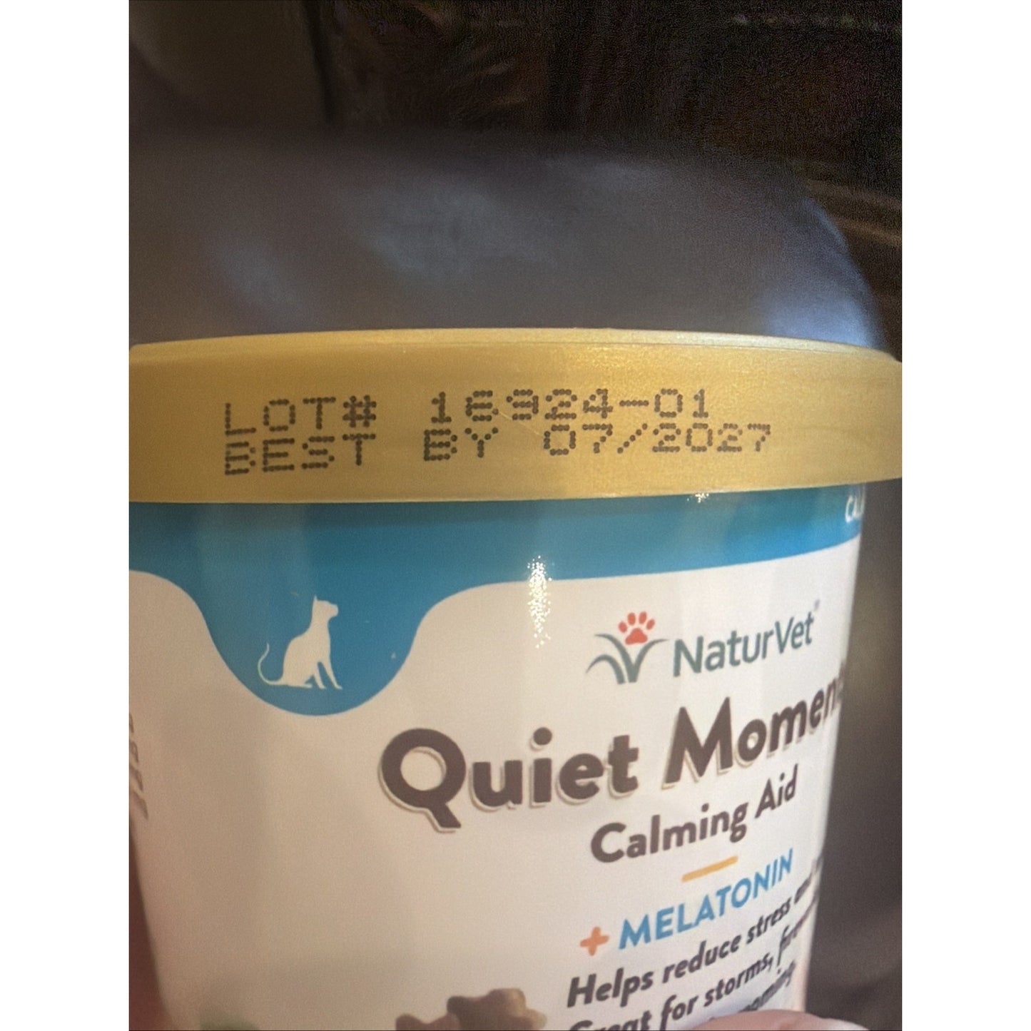 Close-up of a white and blue container of NaturVet Quiet Moments Calming Aid featuring melatonin, with a gold lid stamped with lot number and best by date text.