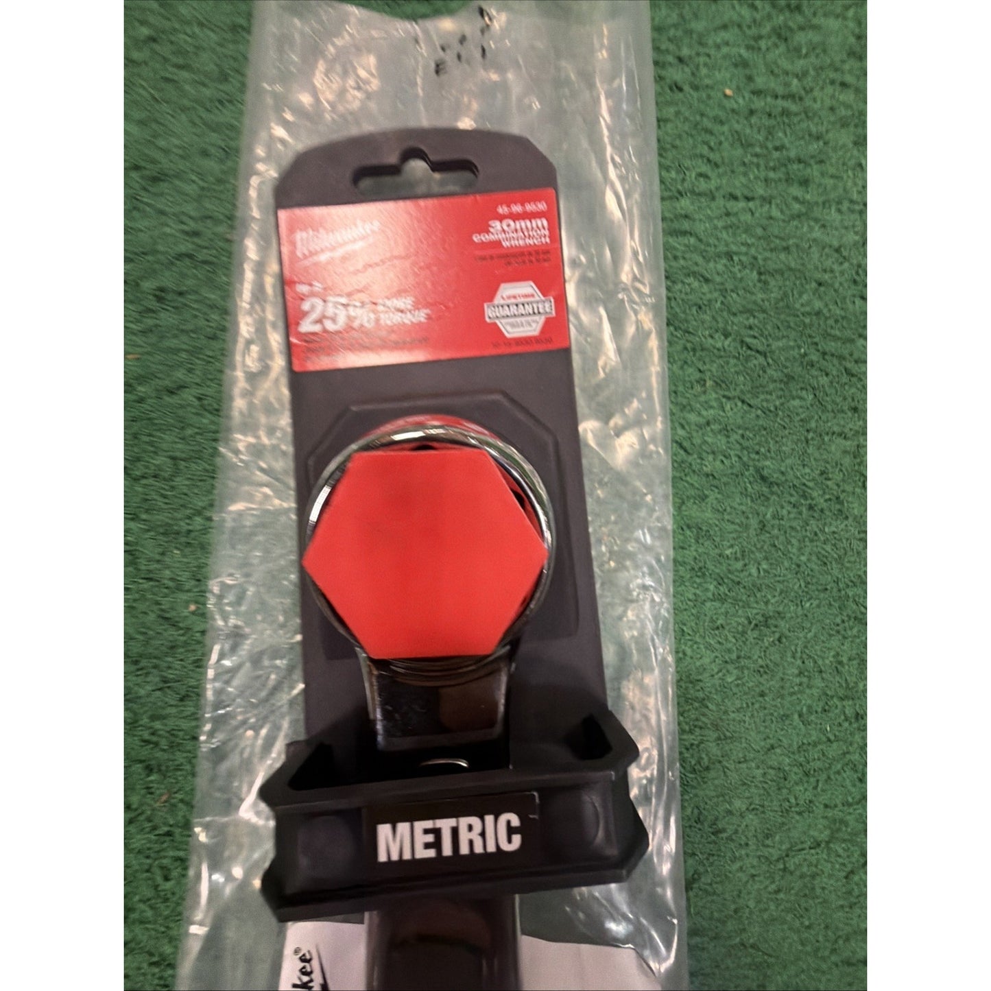 Black plastic wrench grip labeled "METRIC" attached to a chrome-plated hexagonal tool head, displayed on clear plastic packaging with a red and white branded label, set against a green textured surface.