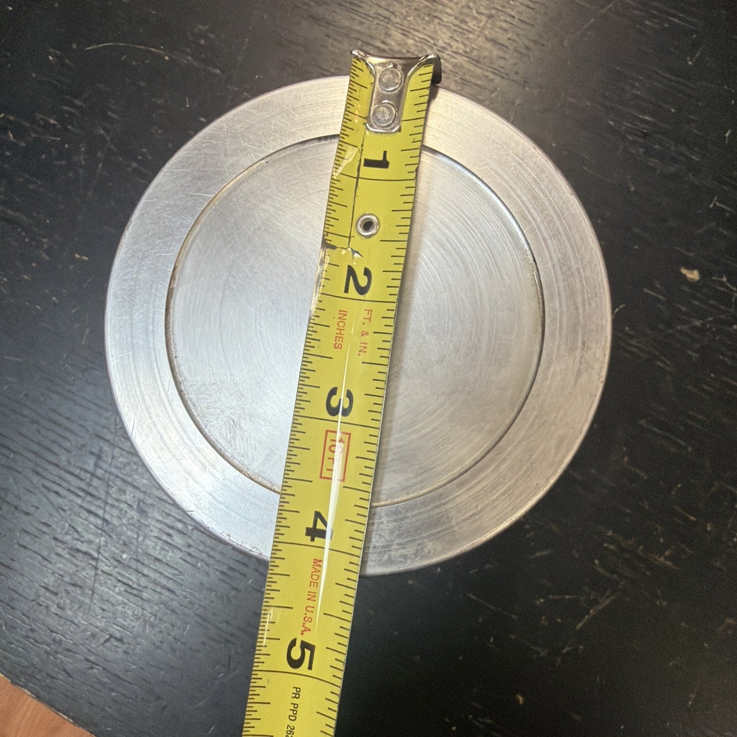 Top-down view of a round metal object with concentric circular grooves, placed on a dark scratched surface, with a yellow measuring tape showing approximately 4 inches in diameter.