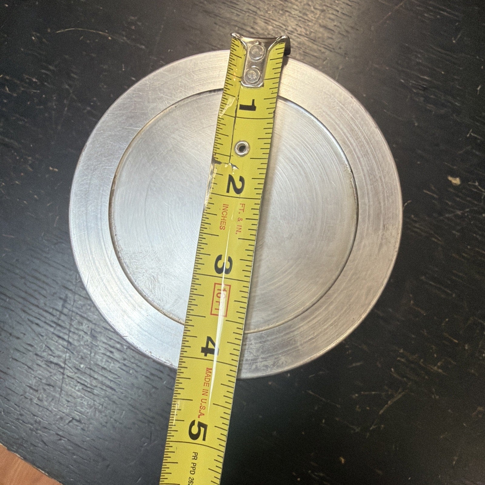 Top-down view of a round metal object with concentric circular grooves, placed on a dark scratched surface, with a yellow measuring tape showing approximately 4 inches in diameter.