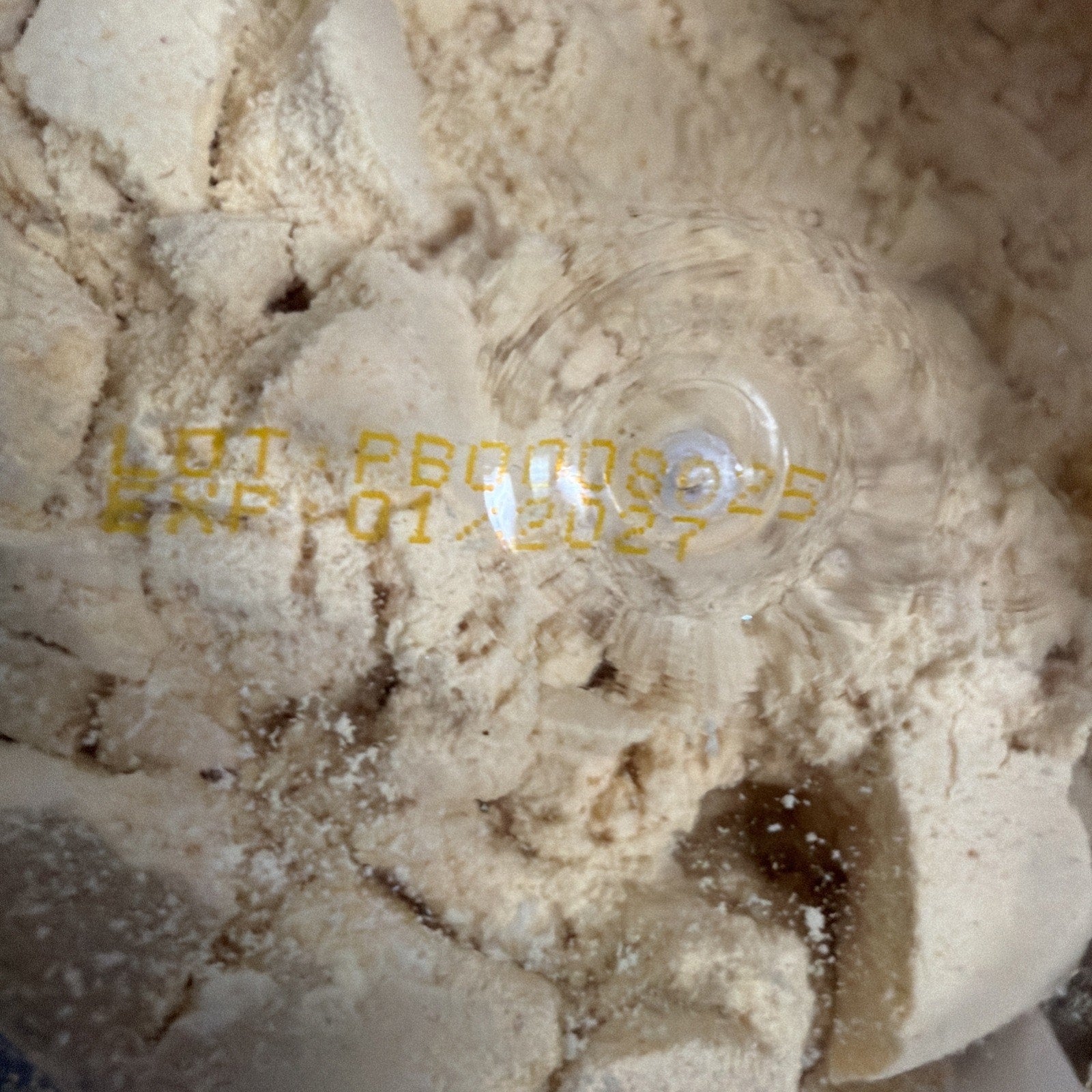 Close-up view of a powdery beige substance with a manufacturing lot number and expiration date printed in yellow ink on the surface beneath a clear plastic covering.