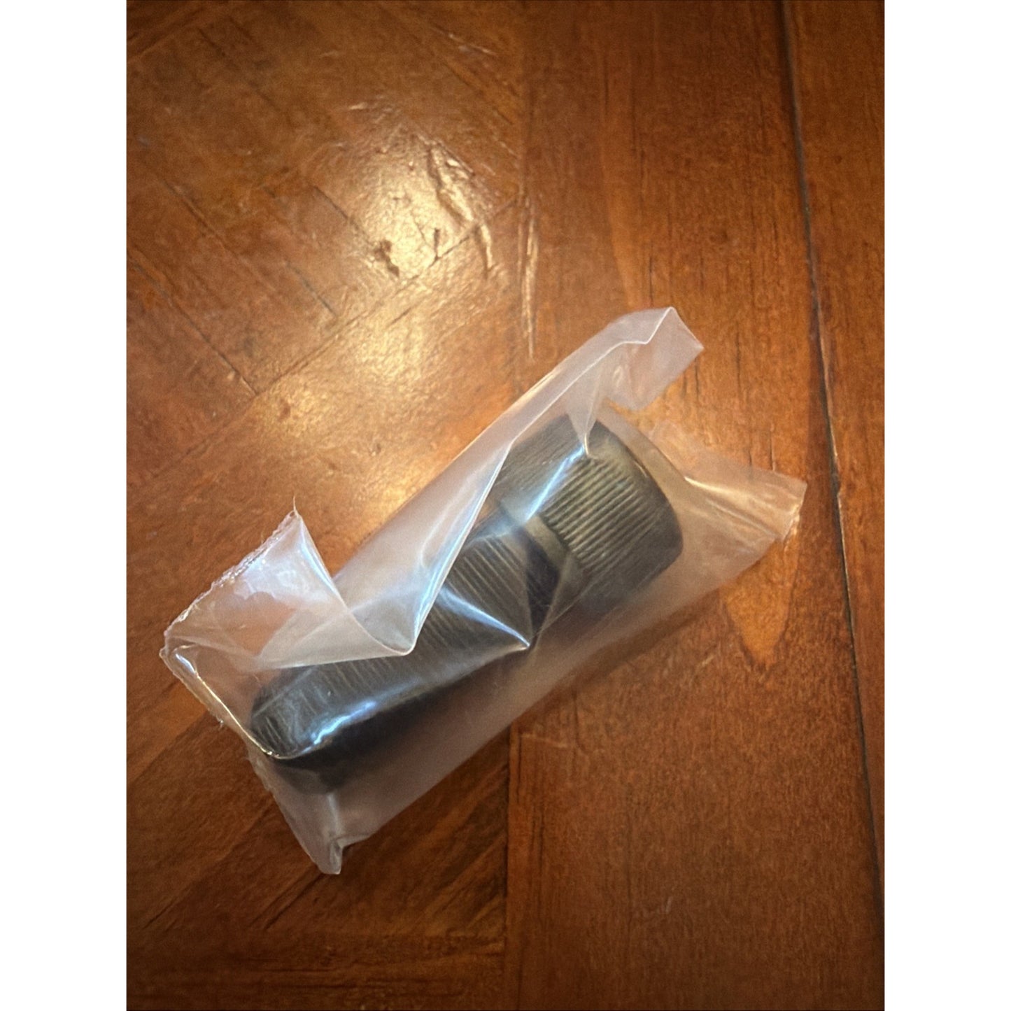 Black cylindrical plastic part with ridged edges inside a clear sealed plastic bag, resting on a wood-grain surface with visible scratches and light reflections.
