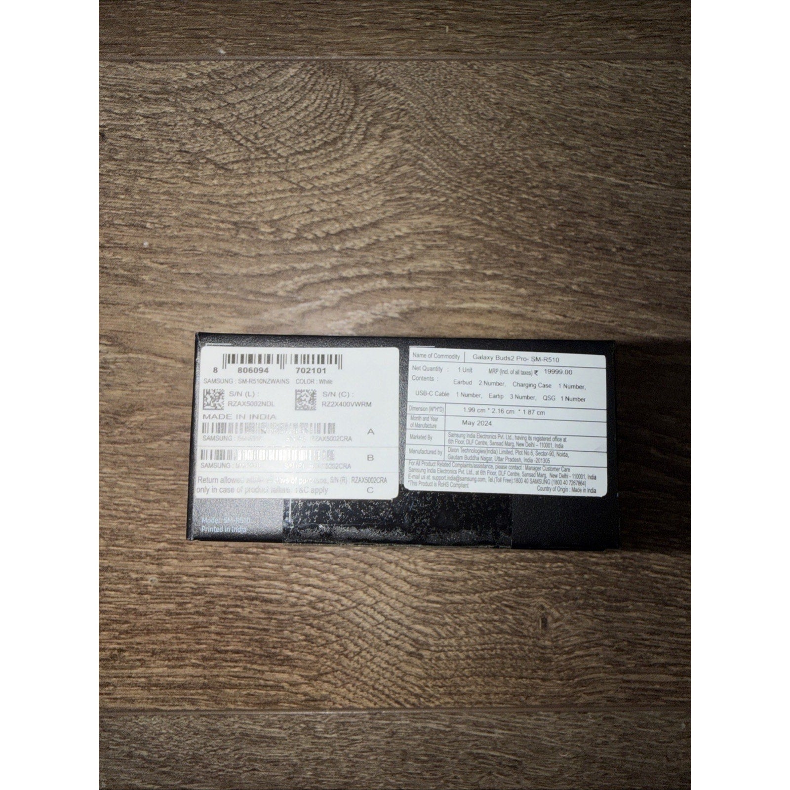 Black rectangular product box displayed on a wooden surface with two white labels containing printed technical specifications, barcode, serial numbers, and manufacturing details.