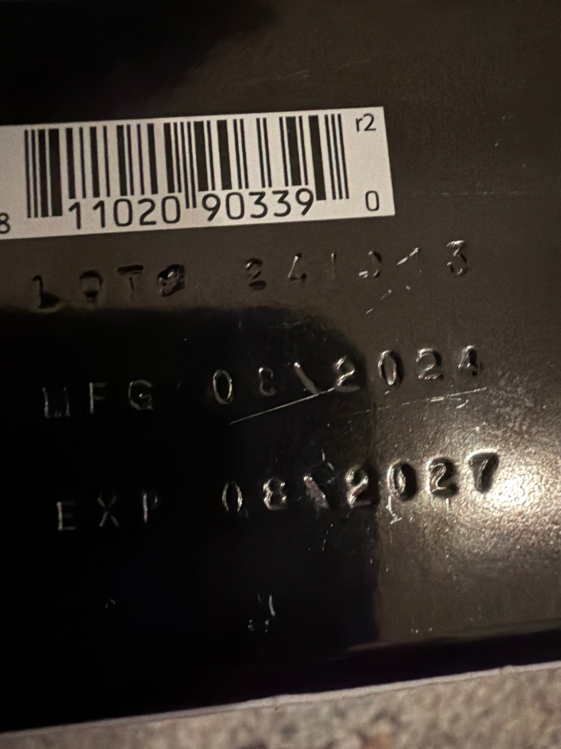 Close-up of a black product surface showing an embossed lot number, manufacturing date of August 2024, expiration date of August 2027, and a barcode sticker with numeric code.