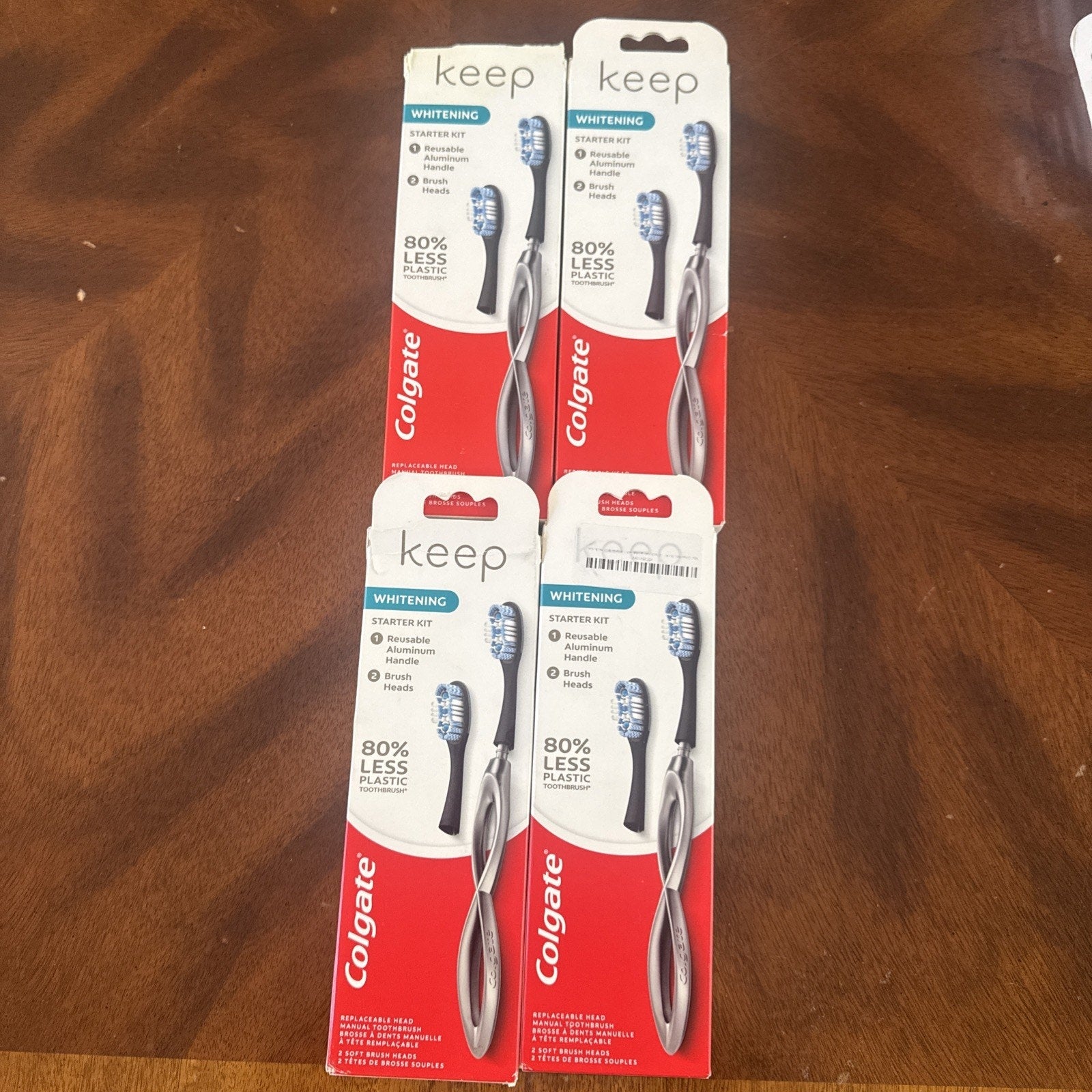 Four packages of Colgate Keep Whitening toothbrush starter kits arranged on a wooden surface, each highlighting 80% less plastic in the brush handles.